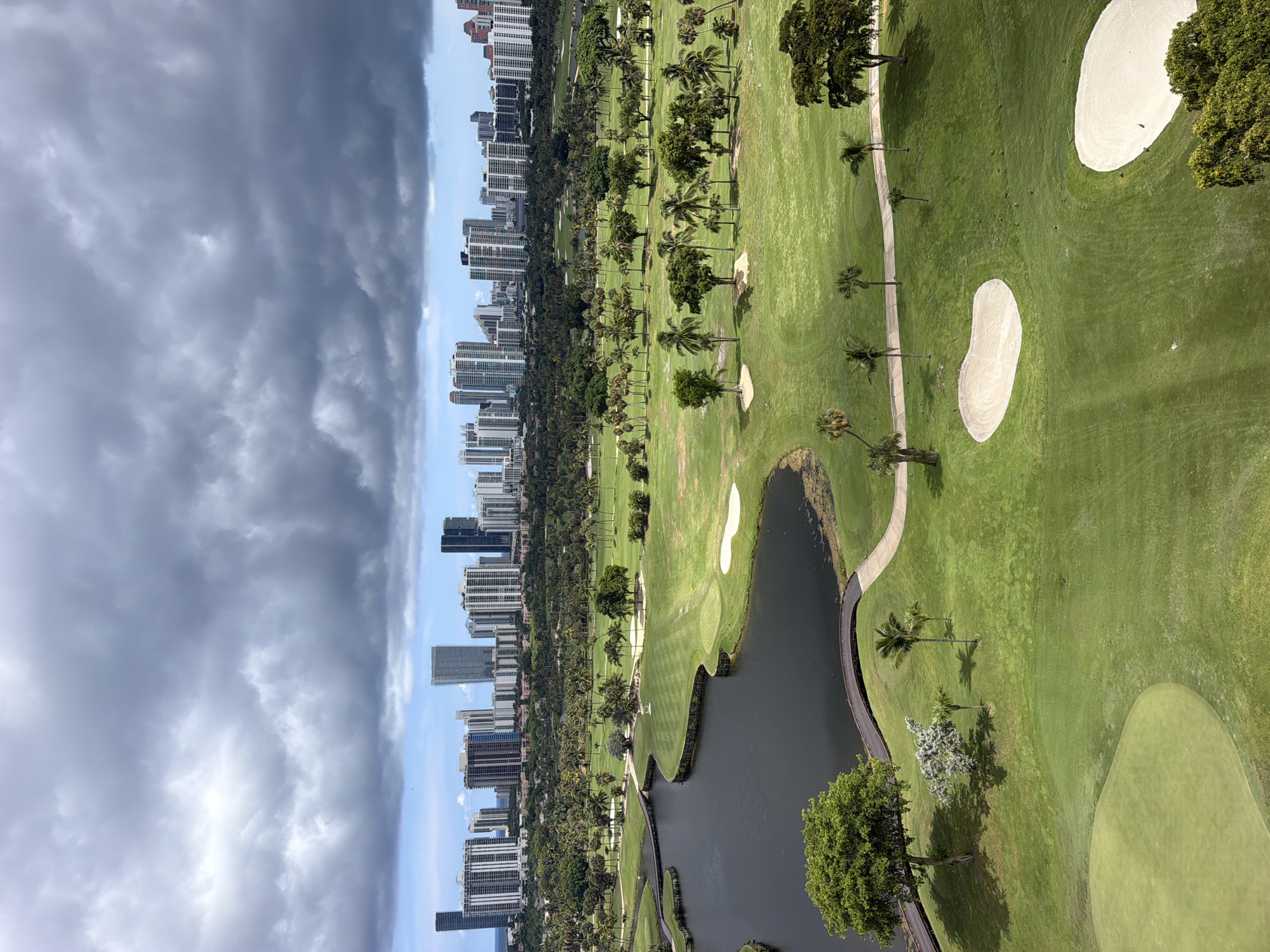 An aerial view captures a golf course in Aventura, Florida, with a cityscape and cloudy sky in the background. The foreground features a meticulously maintained golf course with lush green fairways, sand traps, and a dark body of water. Several palm trees and other vegetation are scattered across the course. A paved pathway winds through the landscape. In the middle ground, a dense line of trees separates the golf course from a city skyline composed of numerous high-rise buildings. The sky above is filled with dramatic, dark gray clouds, contrasting with patches of lighter sky and a hint of blue. The overall impression is of a sprawling urban development encroached upon by a natural landscape, under an overcast sky.