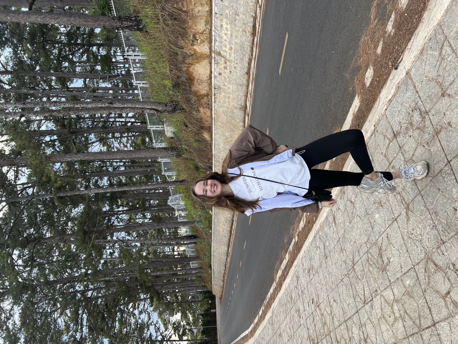 A young female subject stands smiling on a patterned concrete sidewalk, posing with her left knee bent and right hand on her hip. She has light skin, long brown hair, and is wearing a white t-shirt with visible text, an open brown jacket, black leggings, and grey athletic shoes. A small black shoulder bag is visible. The sidewalk borders an asphalt road that features a faint yellow-orange dashed line. Beyond the road, an exposed earth bank with a light-colored retaining wall supports an elevated slope. This slope is densely forested with tall, slender pine trees that extend across the entire background. A white metal fence runs horizontally through the trees on the upper part of the slope. The sky is visible through the tree canopy. This setting is characteristic of the pine-forested roads found within the city of Da Lat, Vietnam.