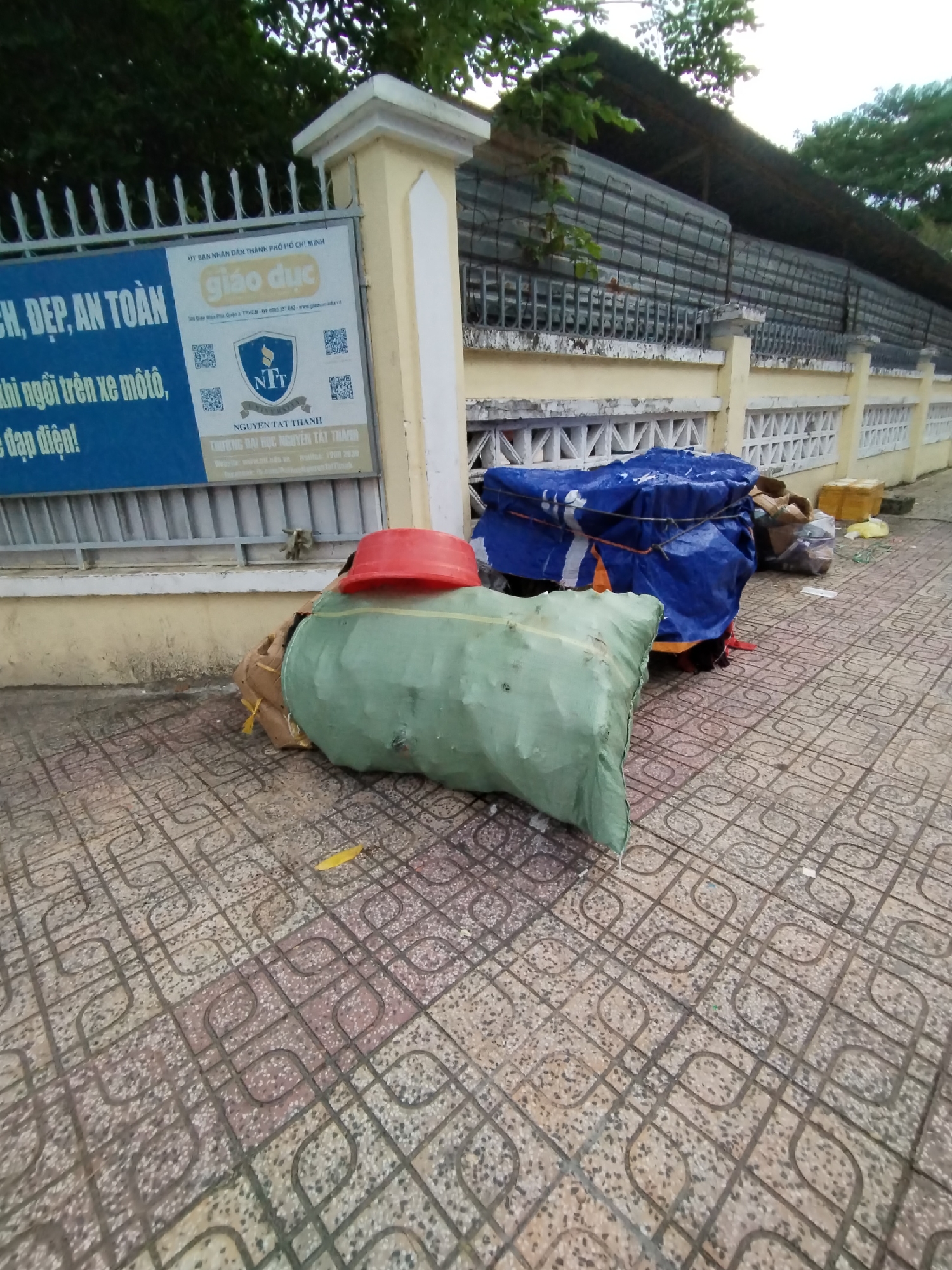 An outdoor, slightly elevated shot captures a section of a sidewalk next to a building or perimeter wall, likely in Bac Nha Trang Ward, Vietnam. In the foreground, several bulky items are left on the patterned, grey and red tiled pavement. A large, rectangular package, wrapped in light green fabric and secured with rope, dominates the left side of the frame, topped by a red plastic basin. To its right, a pile of goods is covered with a blue tarp, also tied down, with cardboard boxes and other items visible beneath and around it. Scattered debris, including a yellow leaf and small pieces of paper, are present on the sidewalk.
Behind these items, a low, light yellow wall or fence with white decorative ventilation blocks runs horizontally. Above this wall is a black metal fence, and in the background, a dark, corrugated roof structure is partially visible, suggesting a building or covered walkway. Green foliage from trees is seen above and behind this structure, indicating an overcast daytime setting with no direct sunlight.
A prominent blue and white sign is affixed to the yellow wall on the left. The sign features Vietnamese text, including "ỦY BAN NHÂN DÂN THÀNH PHỐ HỒ CHÍ MINH" (People's Committee of Ho Chi Minh City) and "giáo dục" (education). Part of a public safety campaign is visible, mentioning "AN TOÀN" (safe/safety) and phrases related to riding motorcycles or electric bikes. The sign also displays the "NTT" logo and identifies "NGUYỄN TẤT THÀNH" and "TRƯỜNG ĐẠI HỌC NGUYỄN TẤT THÀNH" (Nguyen Tat Thanh University), along with a website (www.ntt.edu.vn) and hotline number (1900 2030). No people are visible, and there is no discernible activity taking place, suggesting the items are temporarily stored or left unattended.