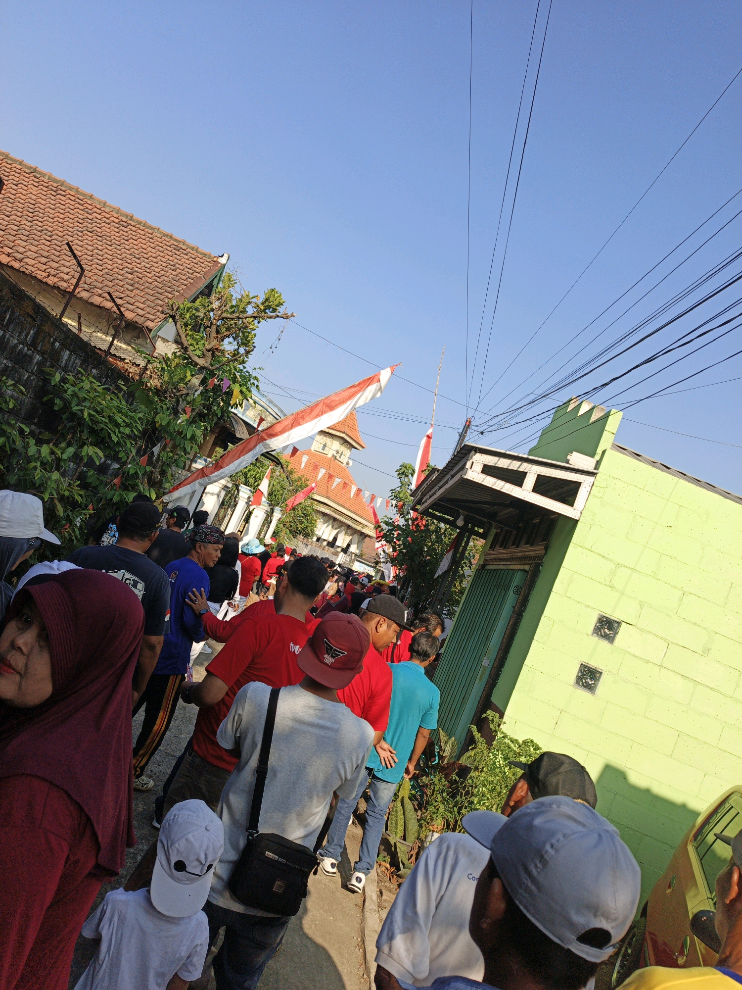 A group of people are walking down a street in Trowulan, Indonesia, during daylight hours. Many of the individuals are wearing red clothing, and Indonesian flags are prominently displayed, suggesting a celebratory or patriotic event. The street is lined with buildings, one of which is painted a bright lime green and features a rustic wooden awning and a green metal door. Another building with a tiled roof and exposed brick is visible in the background. Overhead, a dense network of electrical wires crisscrosses the clear blue sky. The ground surface appears to be a paved road or sidewalk.