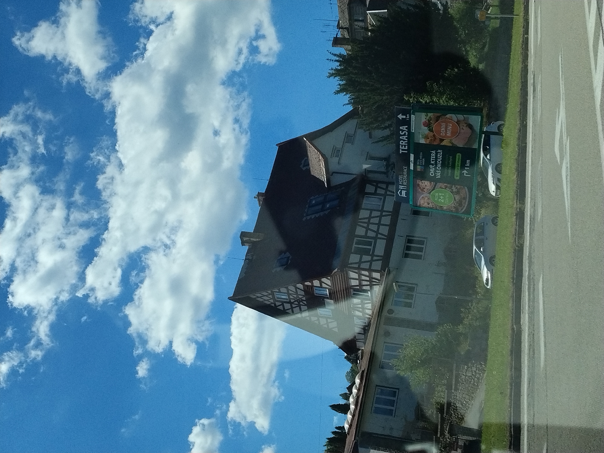 The image shows a view from a vehicle on a sunny day in Vimperk, Czechia. A traditional half-timbered building with a dark tiled roof dominates the right side of the frame. In front of it, a lighter colored building with a flat roof can be seen. A billboard for "HOTEL RESTAURACE TERASA" is prominently displayed, featuring images of food and indicating a distance of 1 km. A white sedan is parked on the grassy verge next to the billboard. The sky is blue with scattered white clouds. The foreground shows a road with painted white lines, indicating a pedestrian crossing. The overall scene depicts a roadside view in a town.