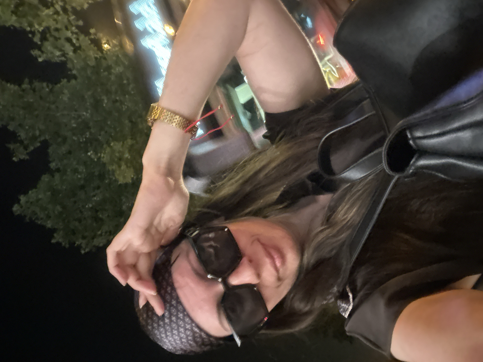 A medium shot selfie shows a woman in a black shirt, large sunglasses, and a patterned headscarf, touching her head. She is wearing a gold watch on her left wrist with a red string bracelet. In the background, blurred lights and trees suggest an outdoor setting at night.