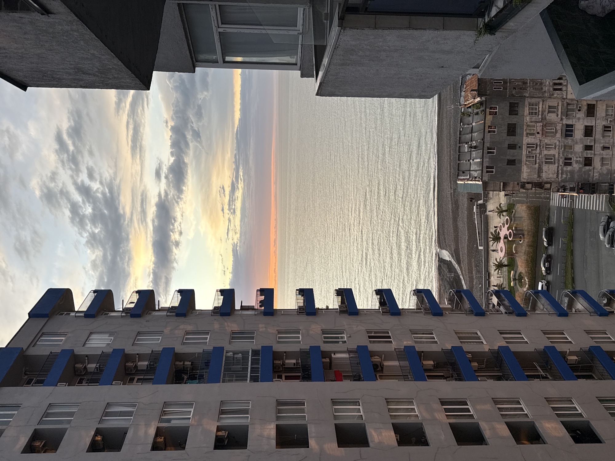 The photograph captures a view from a high vantage point, likely a balcony, showcasing a slice of Batumi, Georgia, juxtaposed against the backdrop of the Black Sea. The most prominent feature is a tall, multi-story residential building with a gray facade and repetitive rows of rectangular windows. Each level is adorned with short, blue-accented balconies, some featuring external air conditioning units and what appears to be ladder-like structures.
To the left, a narrow passage reveals a vivid sunset sky with streaks of orange and yellow light filtering through the clouds above the Black Sea. The sea itself is calm, reflecting the colors of the sunset.
In the far distance, beyond the sea, land is barely visible, merging with the horizon. A sliver of the beach is visible, along with a portion of the coastal road with cars parked and moving.
The overall impression is an urban landscape set against the vast expanse of the sea and sky.