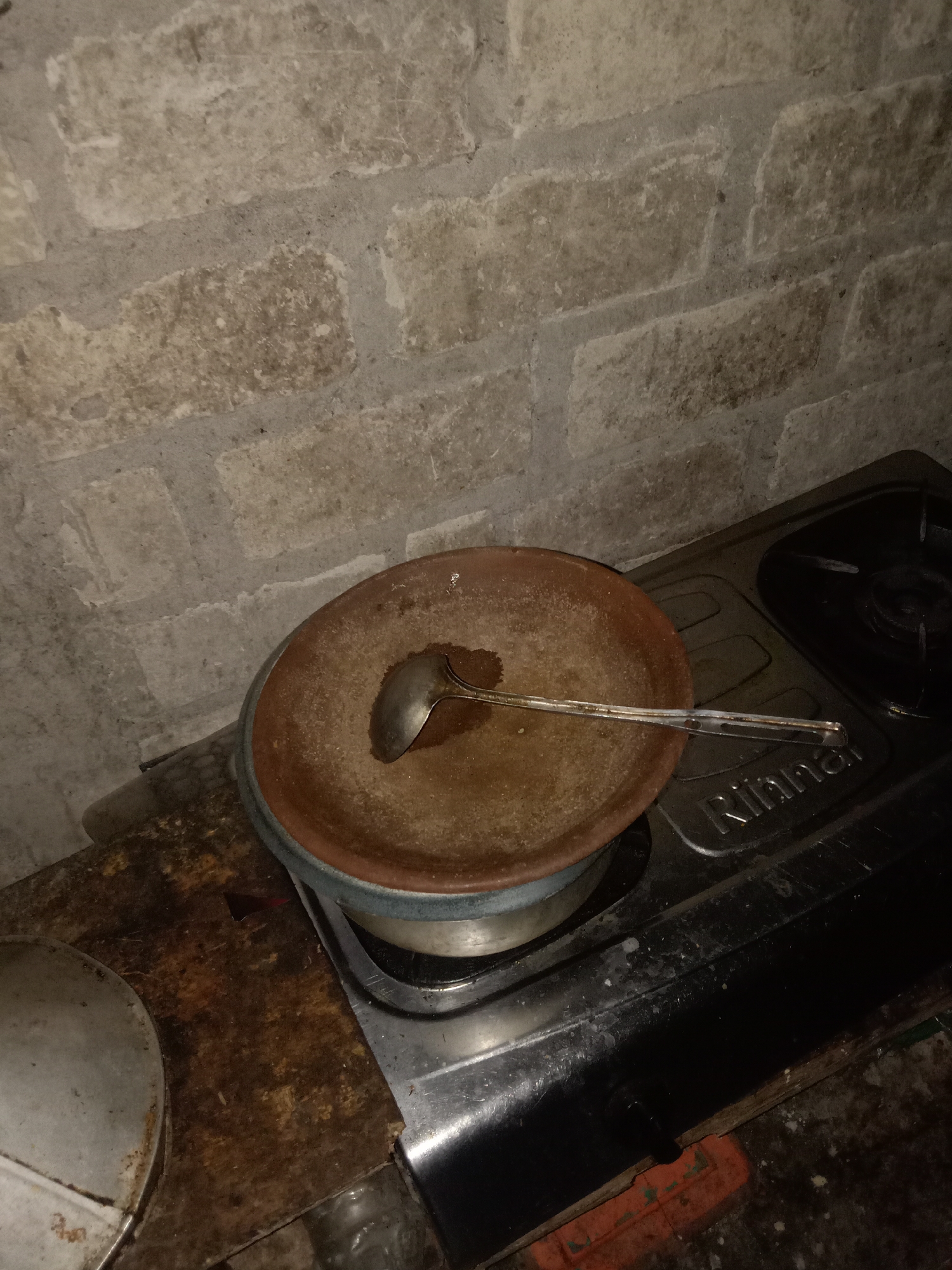 The image shows a kitchen counter in what appears to be a residence in Jumput, Indonesia. On the counter, a clay pan with a ladle sits on top of a gas stove. The wall behind the stove is made of light-colored bricks with visible mortar lines. The counter surface is stained and appears to be made of wood, with some metal elements visible. To the left, a part of a metal pot lid is also present on the counter. The scene suggests a utilitarian kitchen setting.