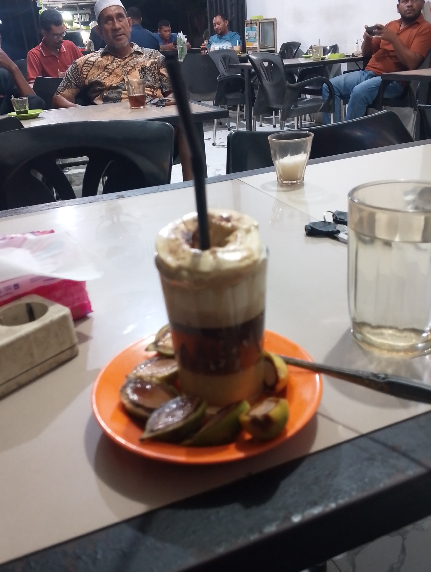 In an outdoor eating area, likely a warung or roadside stall in Cotseutui, Indonesia, several people are seated at tables. In the foreground, a layered beverage in a clear glass with a straw sits on an orange saucer, accompanied by sliced fruit, possibly salak. To the right, a glass of water is visible. In the mid-ground, an older man wearing a white cap and a patterned shirt is seated at a table, looking towards the left. Another man in a red shirt is seated to his left, and a man in a blue shirt is seated further back. The environment appears casual, with simple tables and chairs, and the lighting suggests it is evening or indoors with artificial light. The overall scene depicts people enjoying refreshments in a local eatery.