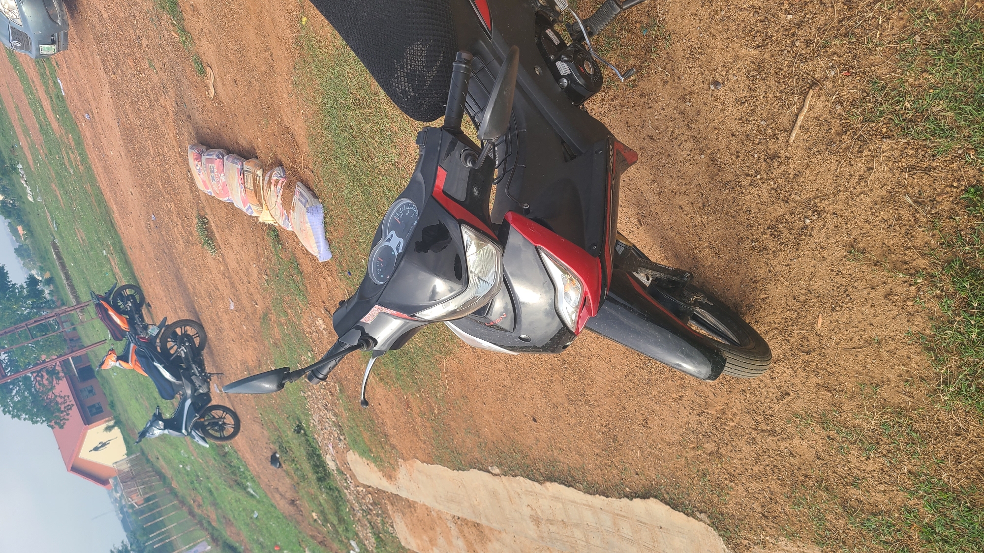 A black and red motorcycle is parked on a brown dirt surface mixed with grass. To the left of the motorcycle, there is a stack of bags. Further in the distance to the left, there are two additional motorcycles parked near a building. The location appears to be outdoors, possibly in Ogbomosho, Nigeria.