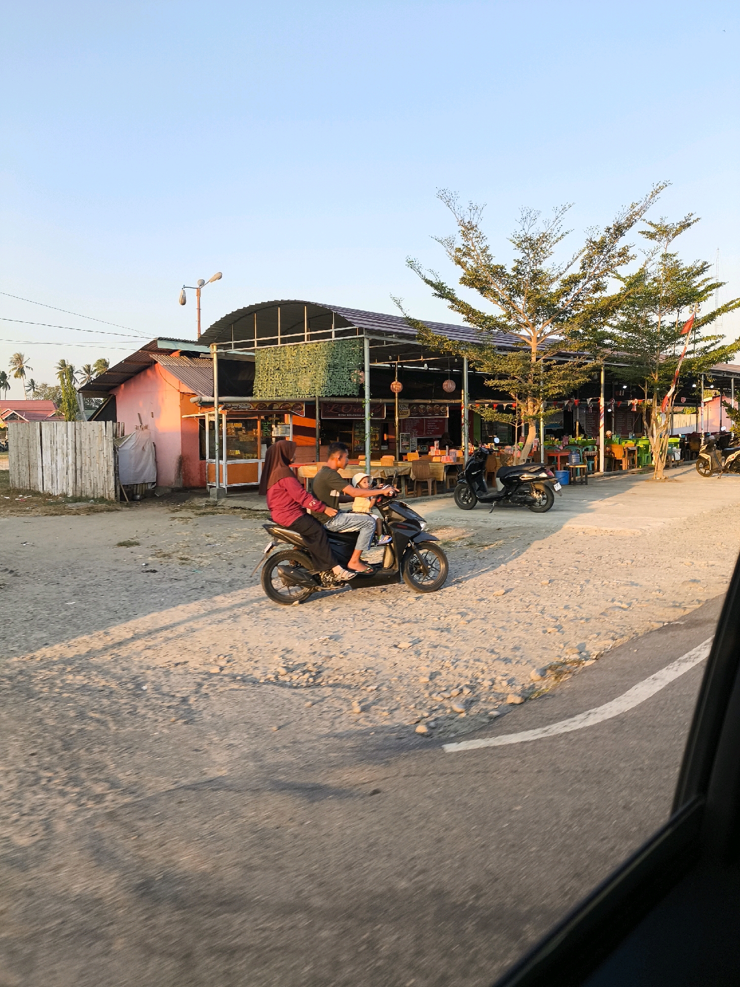 In the city of Tenilo, Indonesia, a snapshot captures a sun-drenched scene. Two people ride a motorcycle, a man driving and a woman with a child passenger, on a dusty road. The woman wears a pink hijab and dark clothing, while the man is in a dark t-shirt and jeans. The child is in a yellow shirt. Behind them, a cluster of buildings suggests a commercial or residential area. One building, painted pink, has a thatched roof and a bamboo fence. Another, larger structure, appears to be a marketplace or restaurant with an open-air seating area filled with tables and chairs. Several trees with sparse, green foliage dot the landscape, and the sky is a clear, pale blue. The overall impression is one of a warm, late afternoon in a developing community.