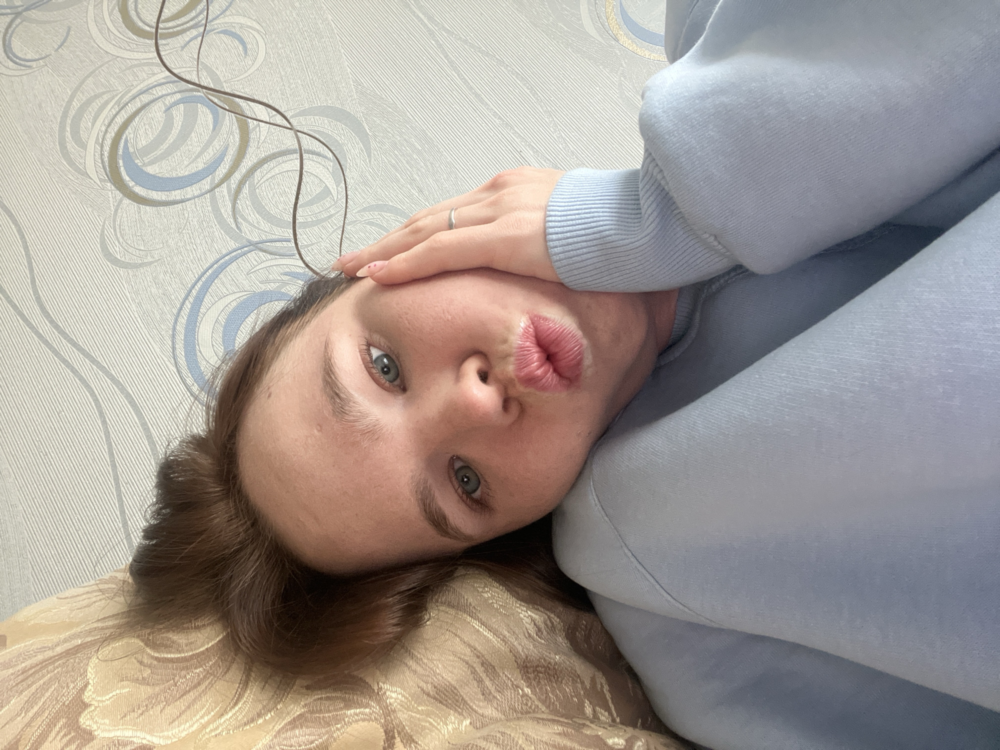 This is a close-up, informal portrait of a young woman, likely a selfie, taken from a slightly tilted, low-angle perspective. The woman is lying down, resting her head on a patterned, light-colored pillow or blanket. She is making a playful "kissing face" with her lips puckered. Her gaze is directed towards the camera with light blue or green eyes.
She has fair skin and light brown hair that frames her face. She is wearing a soft, light blue long-sleeved top, possibly a sweatshirt. Her left hand is resting on her forehead, revealing a silver ring on her ring finger and neatly manicured nails, painted light pink with small decorative dots.
The background features light-toned wallpaper with an abstract, repeating swirl pattern in shades of grey, light blue, and subtle gold or brown. A thin, dark brown cable or wire is visible against the wallpaper in the upper left corner of the frame. The lighting is soft and even, indicating an indoor setting. There is no visible text in the image. The overall mood is casual and relaxed. The location is contextualized as Chaikovsky, Russia.