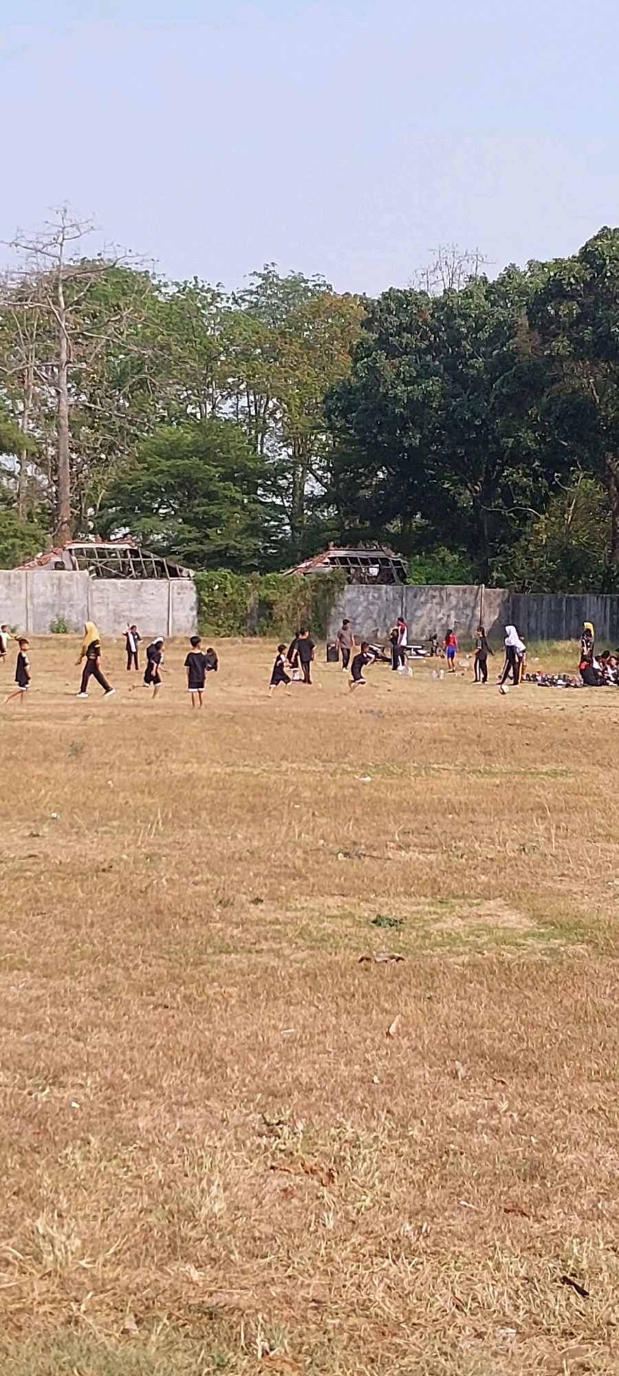 The image depicts an outdoor scene on a dry, grassy field, likely a sports field or open area. In the background, there is a low concrete wall bordering the field, with dilapidated structures behind it, suggesting some form of construction or ruin. Trees form a backdrop behind these structures, with a clear sky above.
The foreground and midground are populated by numerous individuals, predominantly children and young adults, engaged in various activities. Many are wearing dark clothing, possibly uniforms or practice attire, with some wearing yellow accents. Several appear to be participating in some form of organized physical activity or training, with some in motion as if running or performing exercises. There are also some adults present, observing or interacting with the younger individuals.
One notable activity involves a group of individuals wearing black outfits with yellow headscarves, suggesting a martial arts or dance practice. Some are in stances or mid-movement. Another group, further to the right, appears to be gathered near some bottles and possibly a mat, with some individuals in athletic wear.
The overall impression is a community gathering or training session taking place in a somewhat rustic outdoor environment. The location is identified as Keling, Indonesia, which suggests a tropical or subtropical climate given the dry grass and lush trees.