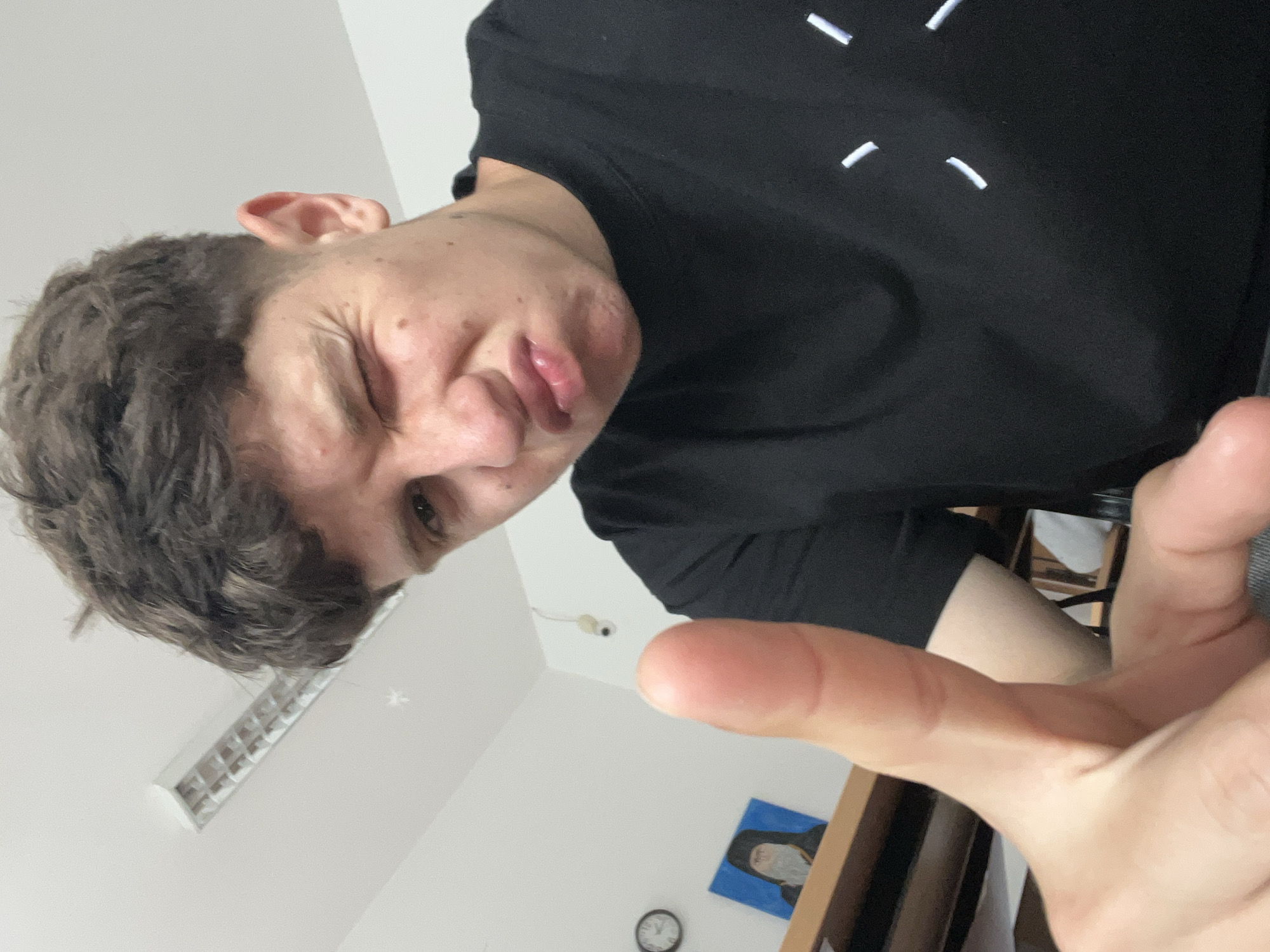 A young male individual is captured in a close-up, low-angle indoor photograph, possibly a selfie, within a room in Zlatograd, Bulgaria. He is looking directly at the camera with a playful and exaggerated facial expression, featuring a crinkled face, wide visible eye, and puckered lips. He has short, curly dark brown hair and is wearing a black t-shirt embellished with multiple small, white dashed lines arranged in a circular pattern on the front.
His right hand is extended towards the camera, with the thumb and index finger slightly raised, creating a sense of direct interaction or a framing gesture. The background reveals a simple setting with white walls and a white ceiling. Visible overhead is a rectangular fluorescent light fixture. On the wall, there's a small, white ball-shaped device, which appears to be a security camera or sensor. Below it, a small framed picture displays a figure in a dark, hooded robe against a light blue background, alongside a partially visible round wall clock. A section of wooden furniture is also seen in the bottom right. The room is lit by artificial light.