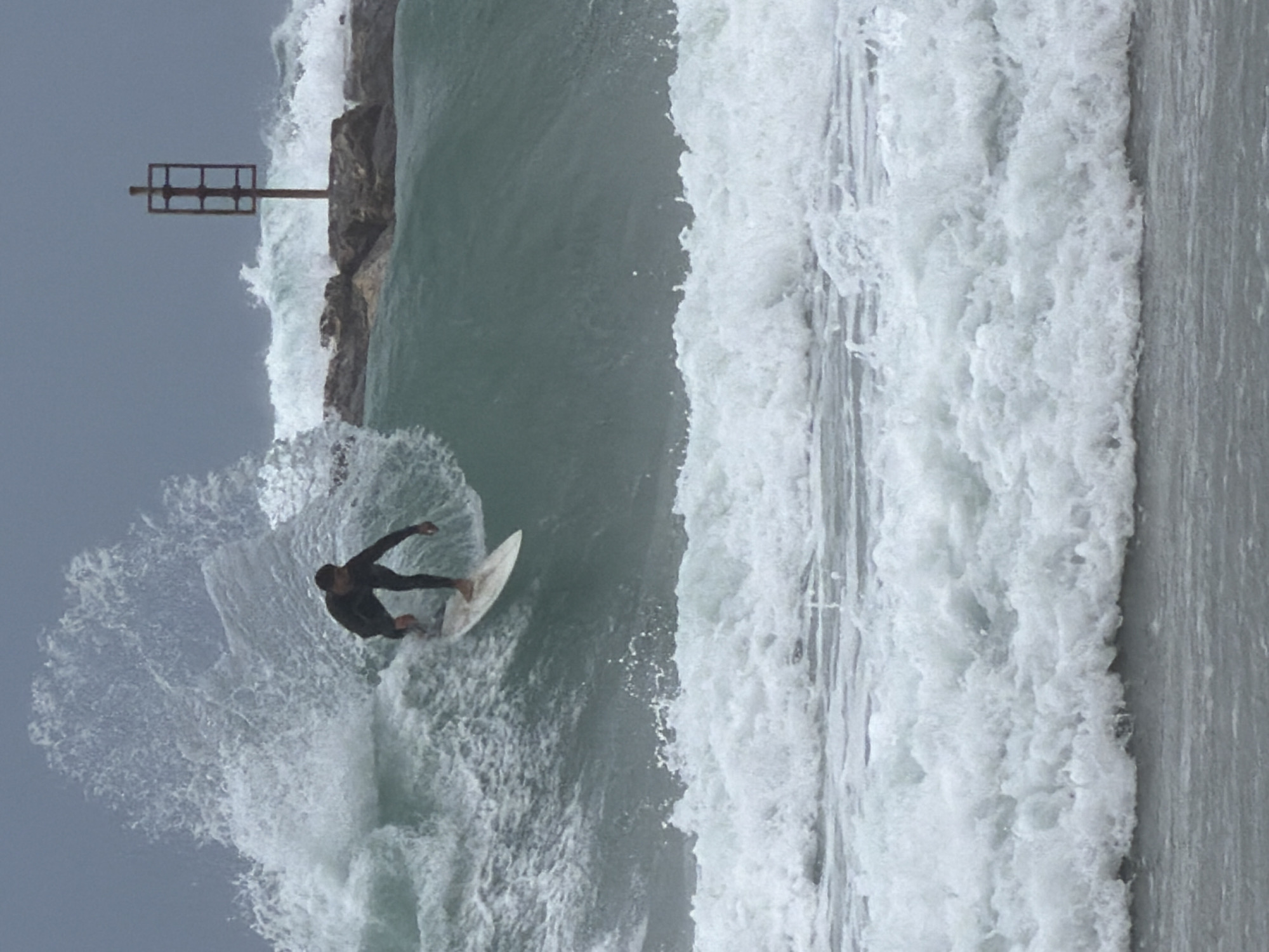 A surfer in a wetsuit rides a wave adjacent to a rocky breakwater with a metal structure, likely in Tel Aviv, Israel. The sea is turbulent with breaking waves and white water. The sky is overcast.