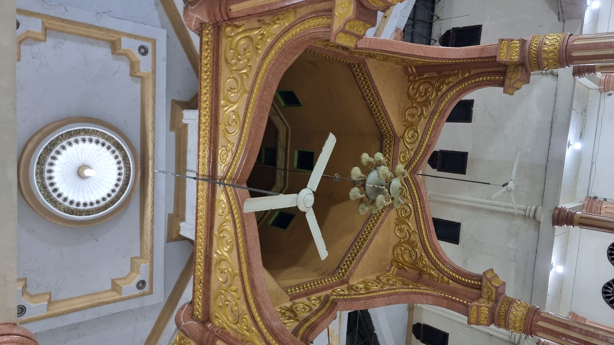 The image shows the interior of a building, likely a mosque, with ornate architectural details. A ceiling fan is suspended from above. The ceiling features a circular design with decorative molding. The walls are white and have dark windows. There are also decorative arches and columns visible. The location is specified as Manetunong, Indonesia.