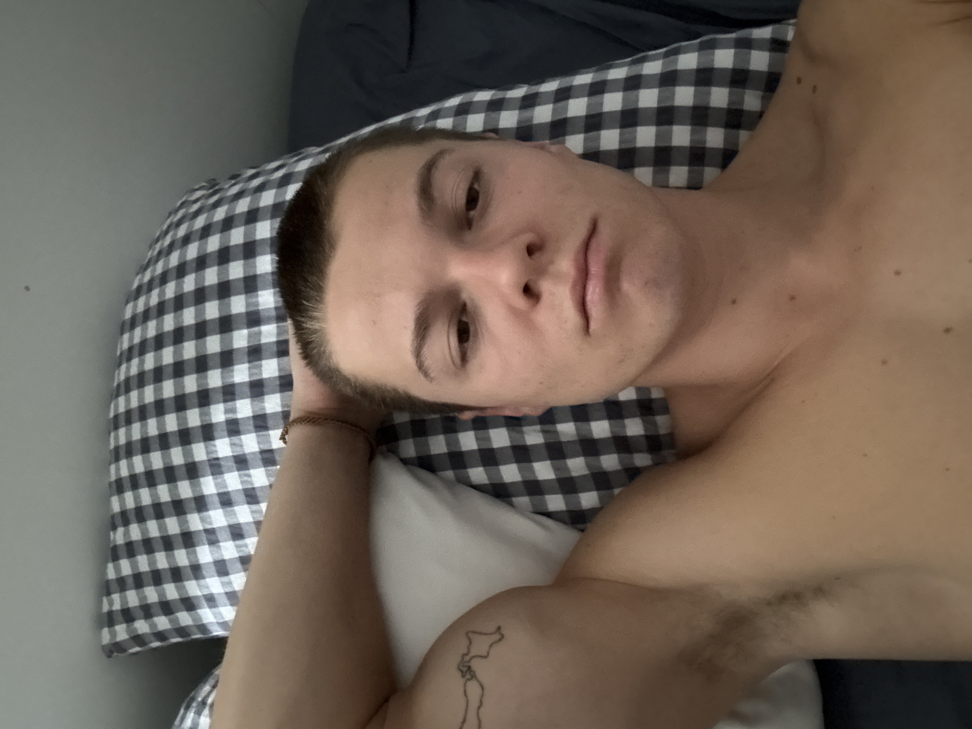 A fair-skinned man with short brown hair lies on his back in a bed. He is shirtless. His left arm is bent at the elbow and his hand is behind his head. The pillow beneath his head has a black and white checkered pattern. A small tattoo is visible on his upper right arm. The bedsheets are navy blue. The background appears to be a light-colored wall. The location is not discernible as London, United Kingdom based on this image.