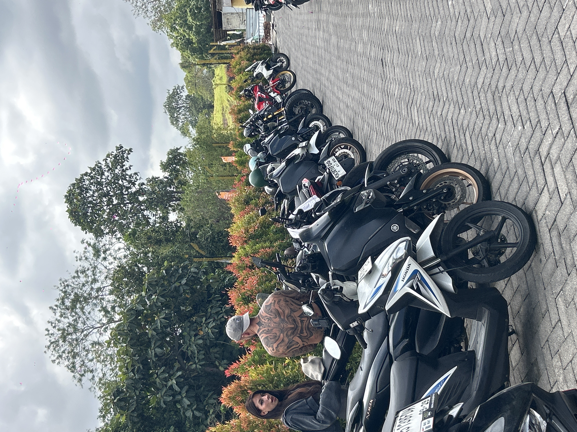 In Mengwi, Indonesia, a lineup of motorcycles is parked on a brick-paved surface. A woman with long dark hair and a grey jacket is seated on the left, looking towards the camera. Beside her, a man with a large, intricate tattoo covering his back and wearing a grey baseball cap is standing. The background features lush green trees and a cloudy sky, with some pink objects scattered in the sky, possibly kites or decorations. The motorcycles are of various models, including scooters and sport bikes, with some helmets visible on the seats. The scene appears to be an outdoor parking area, possibly near a tourist attraction or a rental service.