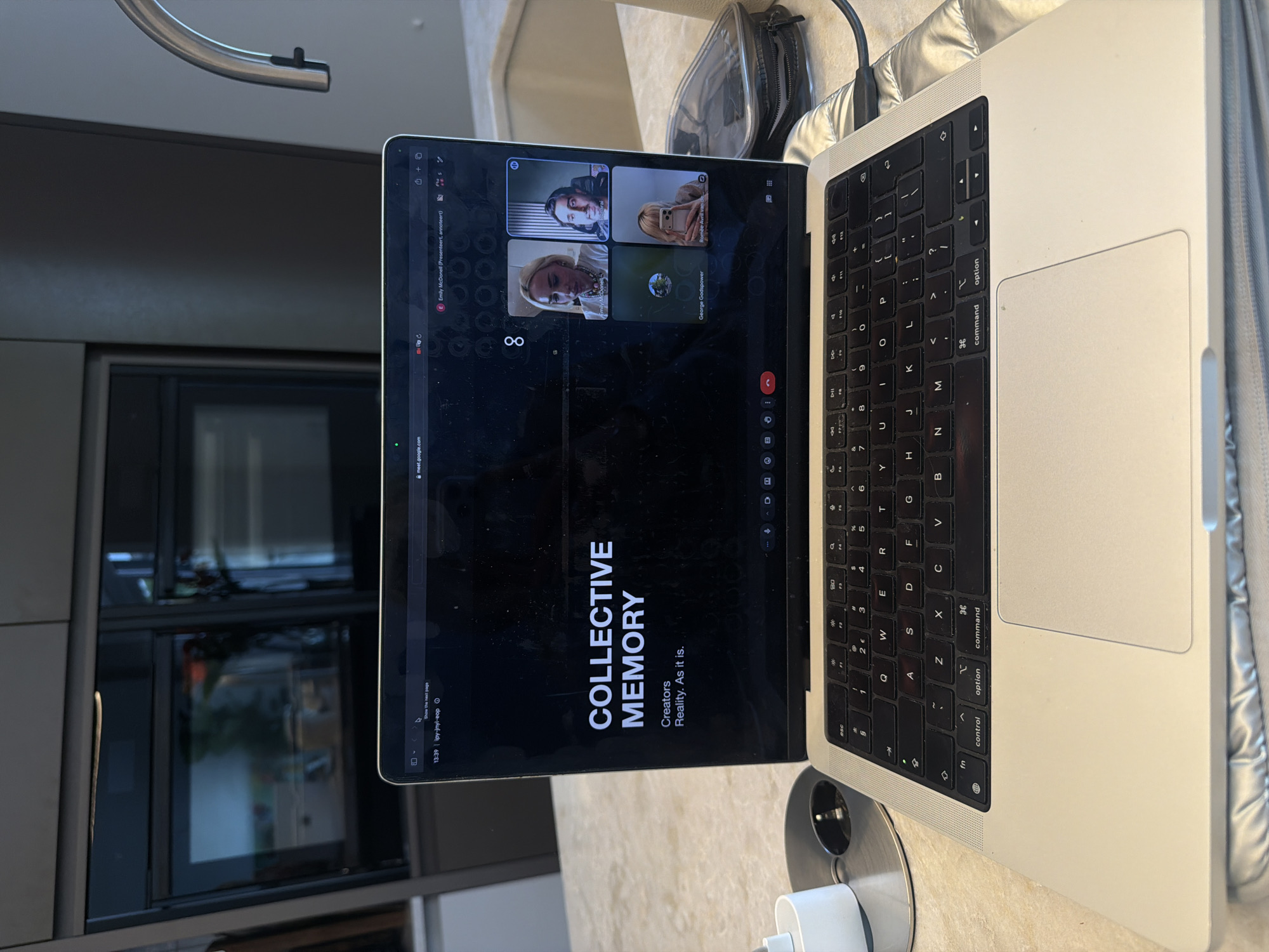 The image shows a close-up view of a laptop open on a countertop, likely in a kitchen. The laptop displays a Google Meet video conference with four participants. The title on the screen reads "COLLECTIVE MEMORY" with the subtitle "Creators Reality. As it is.". The participants are Emily McDonnell, Morris Boharal, George Godspower, and Romée Avril van de Wiel. The background reveals modern kitchen cabinetry with built-in appliances. The scene is set indoors, on a speckled countertop, with a metallic silver laptop as the central subject. The lighting suggests it is daytime, with natural light illuminating the scene. There is no visible indication of the location being specifically in Ridderkerk, Netherlands.