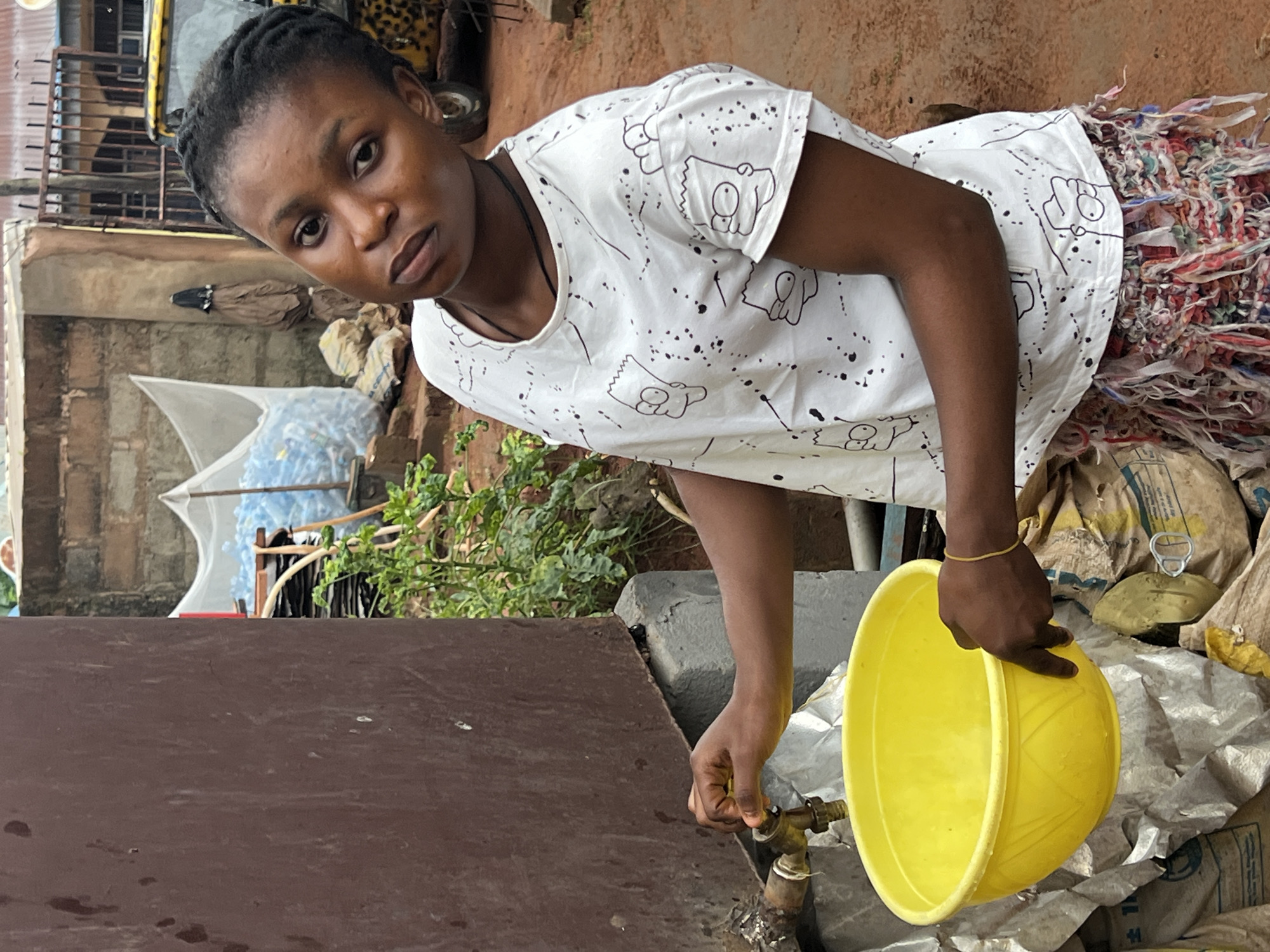 A young woman stands outside a building in Nkpor, Nigeria, collecting water. She is wearing a white t-shirt with a Bart Simpson print and a colorful skirt. Her left hand is on a tap, from which water is flowing into a yellow basin held by her right hand. In the background, there are brick buildings, plastic water bottles, sacks, and a yellow vehicle. The ground is covered in reddish-brown earth.
