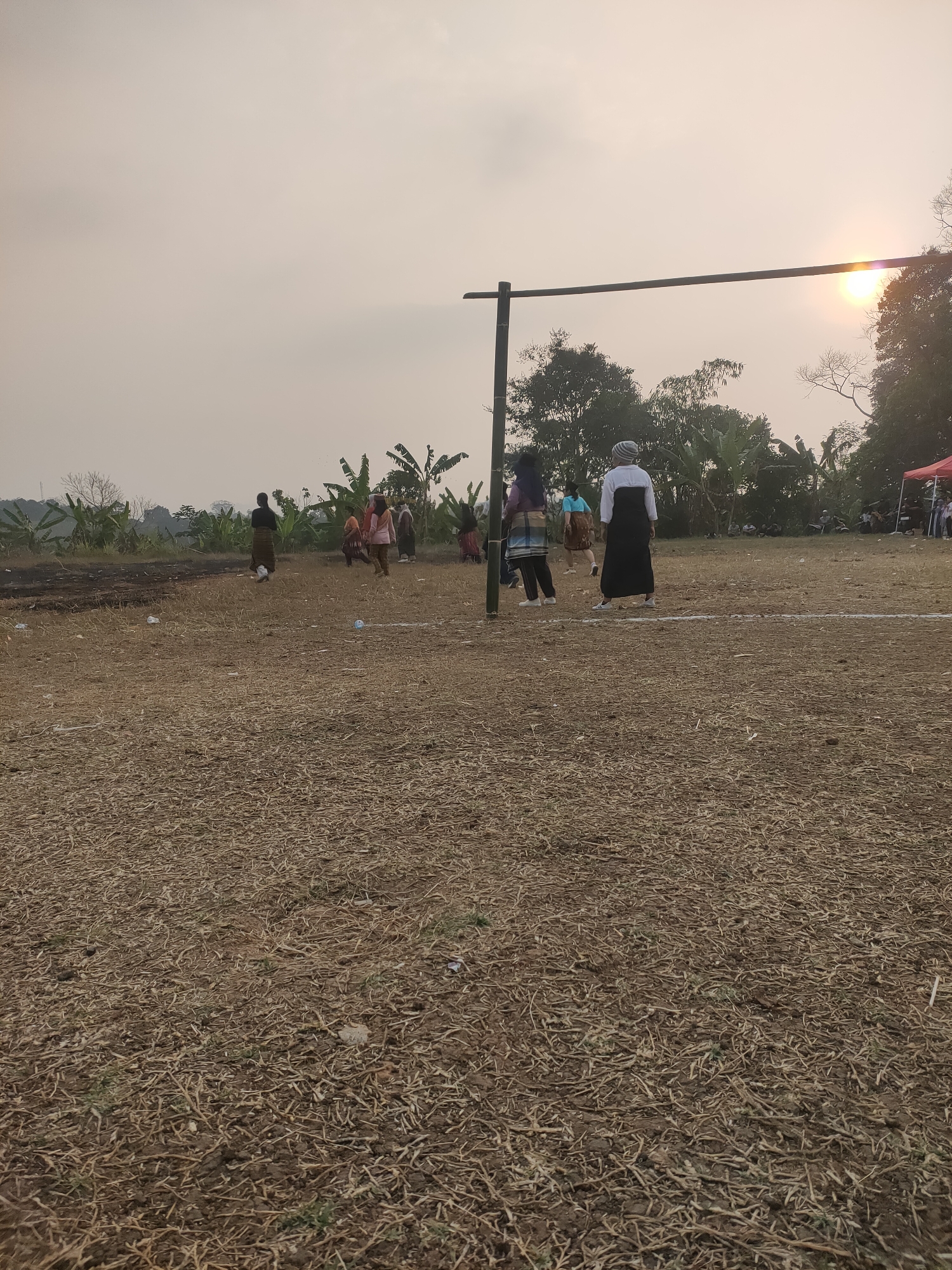 This image captures an outdoor scene in Subang, Indonesia, featuring a group of people gathered on a dry, grassy field. The setting appears to be a sports or recreational area, indicated by a horizontal wooden bar supported by vertical poles, possibly part of a volleyball net setup.
Several individuals are visible in the mid-ground, engaged in various activities. One person, dressed in a white shirt and a black sarong-like garment, stands facing away from the camera. Another person, in a purple and blue patterned skirt and top, is also seen from the back. Other figures in the distance are less distinct but suggest a gathering of people, some appearing to be playing or spectating.
The environment is characterized by dry, short grass and scattered debris, with a smattering of plastic waste visible on the ground. Banana trees and other vegetation form a backdrop, with a dense tree line in the distance. The sky is overcast with a hazy quality, and the sun is visible as a bright, diffused orb, suggesting it might be late afternoon or early morning. In the far right background, a red tent and additional people are present, hinting at a larger event or setup. The overall impression is of a casual community gathering in a rural or semi-rural outdoor space.