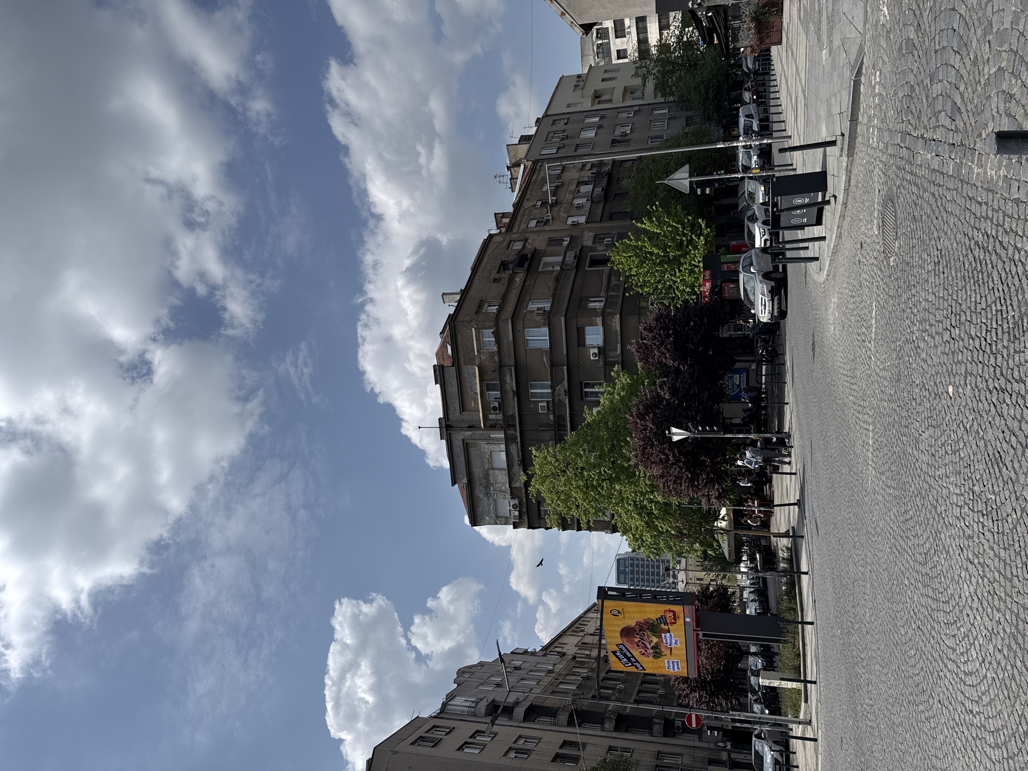 The image depicts an urban street scene on a bright, sunny day with a blue sky and numerous white clouds. The original image is rotated 90 degrees counter-clockwise; when viewed correctly, it shows a street lined with multi-story buildings, typical of an older European city.
On the left side of the street, several aged, grey buildings with numerous windows and visible air conditioning units rise towards the sky. Some buildings have balconies, and antennas are visible on their rooftops. Green trees with lush foliage, along with some trees featuring reddish-purple leaves, are planted along the sidewalk, partially obscuring the lower parts of the buildings.
The street itself is paved with intricate cobblestones and features a paved sidewalk area. Several cars are parked along the street, and in the distance, faint figures of people can be seen, some possibly sitting at outdoor cafe tables. Street furniture includes black, cylindrical trash cans and lampposts. A traffic sign with an arrow pointing right is visible, as is a red circular sign with a white horizontal bar, indicating "No Entry" or "Do Not Enter." A manhole cover is also visible on the cobblestone surface.
On the right side of the street, a large, illuminated billboard stands out. The billboard advertises "Jafa!" and "Plazma, keks i tortice," featuring images of various sweets, including Jafa cakes and Plazma biscuits.
The weather is clear and sunny, casting distinct shadows, suggesting it is mid-day. A small black bird is visible flying in the sky between the buildings. The architecture and visible text cues align with the provided location context of Belgrade, Serbia.