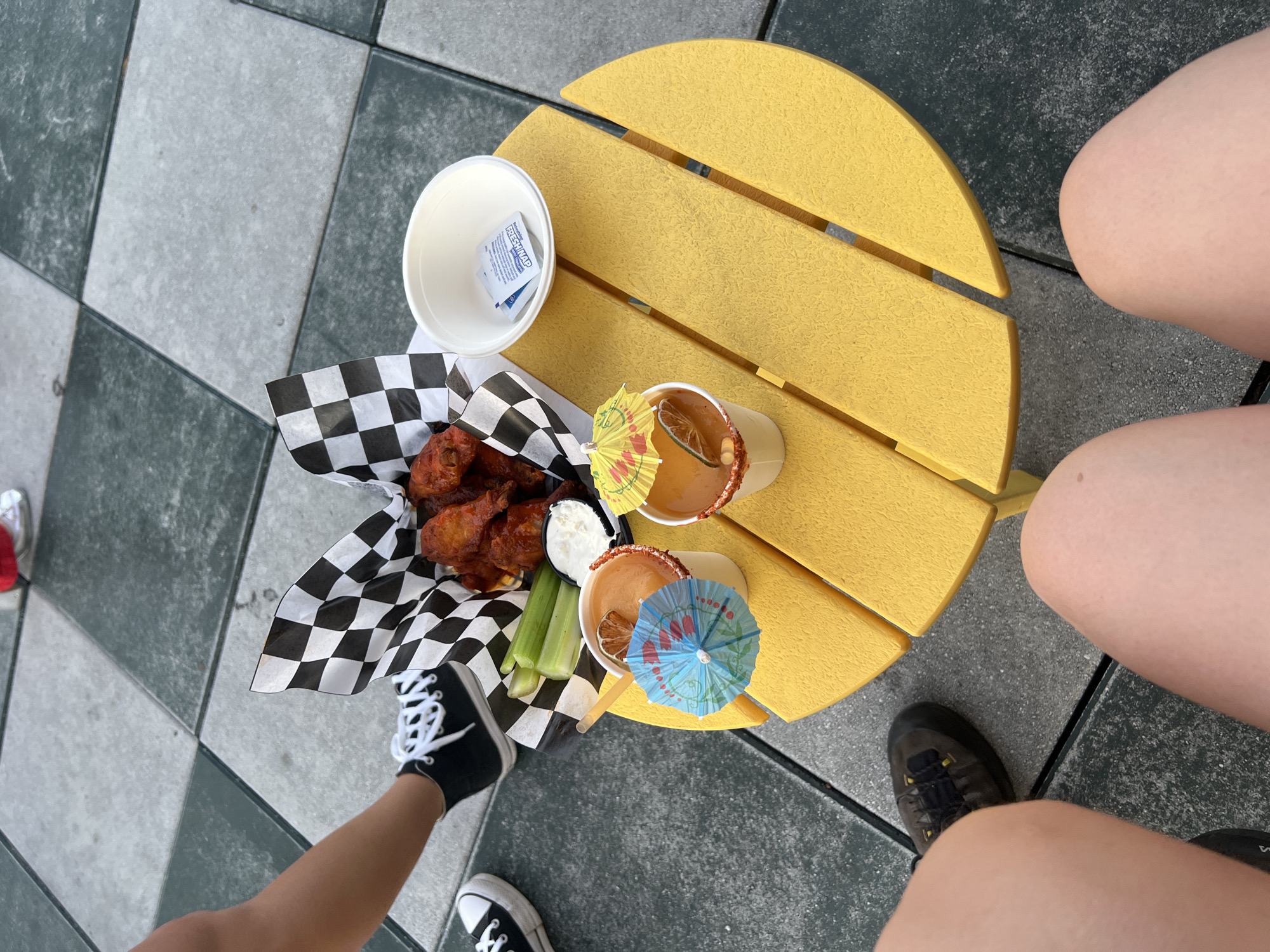 The image depicts a round, yellow wooden table with food and drinks on it, set against a backdrop of a black and gray tiled surface. On the table, a black and white checkered paper tray holds what appears to be chicken wings, celery sticks, and a small container of dip. Next to this, there's a white paper cup filled with individually wrapped hand wipes. Two beverages are also on the table, each adorned with a small paper umbrella and a lime slice. One leg and foot of an individual are visible, wearing a black and white sneaker. Another individual's legs and feet, wearing a dark-colored shoe, are positioned next to the table. The overall scene appears to be an outdoor dining setting, likely in New York City, given the style of tiled surface.