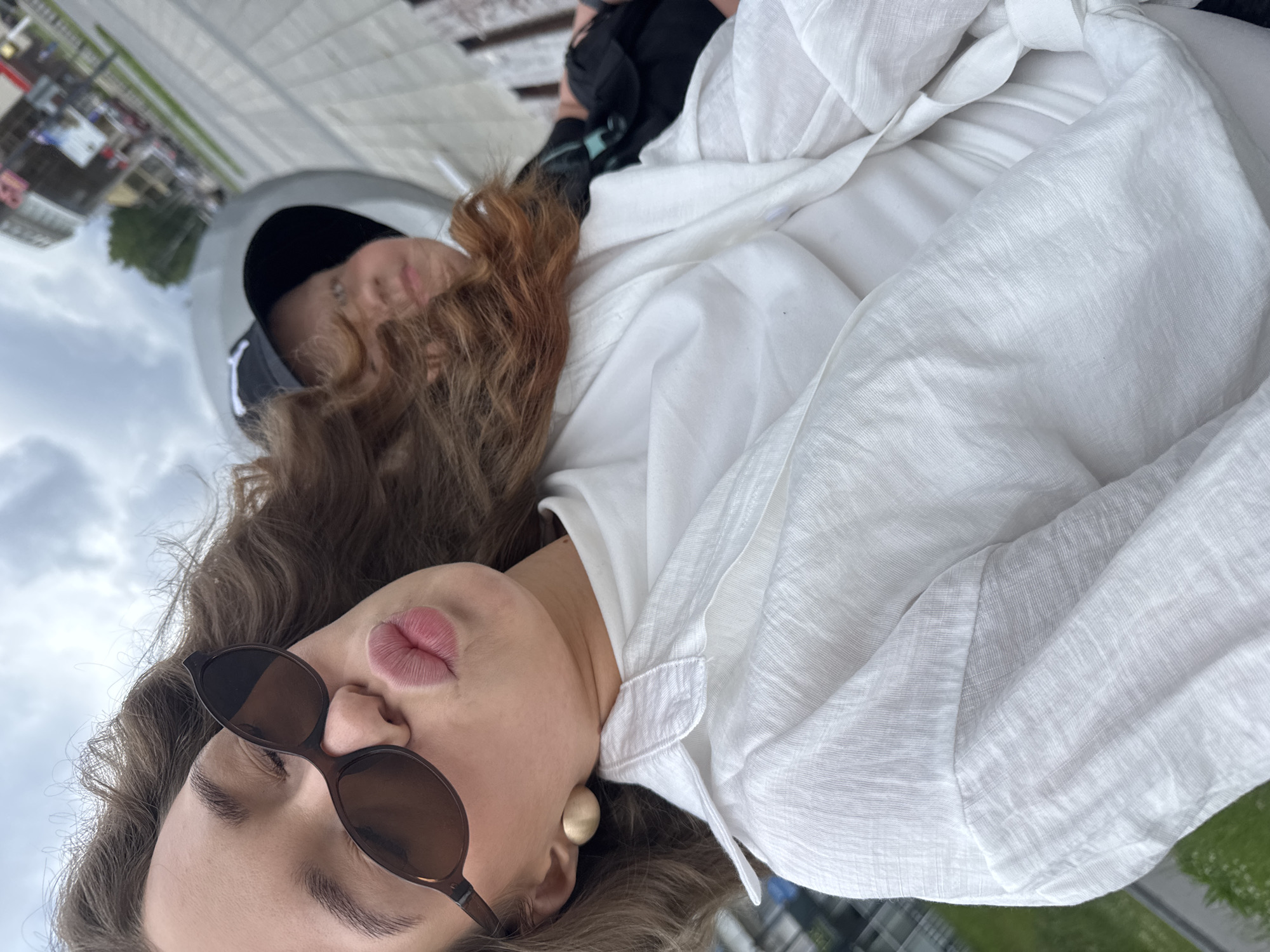 In an outdoor, urban environment, likely in the city of Chaikovsky, Russia, two individuals are captured in a close-up selfie. The foreground is dominated by a young woman wearing large, dark-rimmed sunglasses and a white button-down shirt. She is leaning in, puckering her lips as if for a kiss, and her auburn hair is windblown. A large, gold, circular earring is visible on her left ear. Behind her and slightly out of focus, a second person, possibly a child, is wearing a black baseball cap and a dark garment. They are partially obscured by the woman and her hair. The background shows a paved walkway, green grass, and buildings in the distance, suggesting a public space like a park or promenade. The sky is overcast.