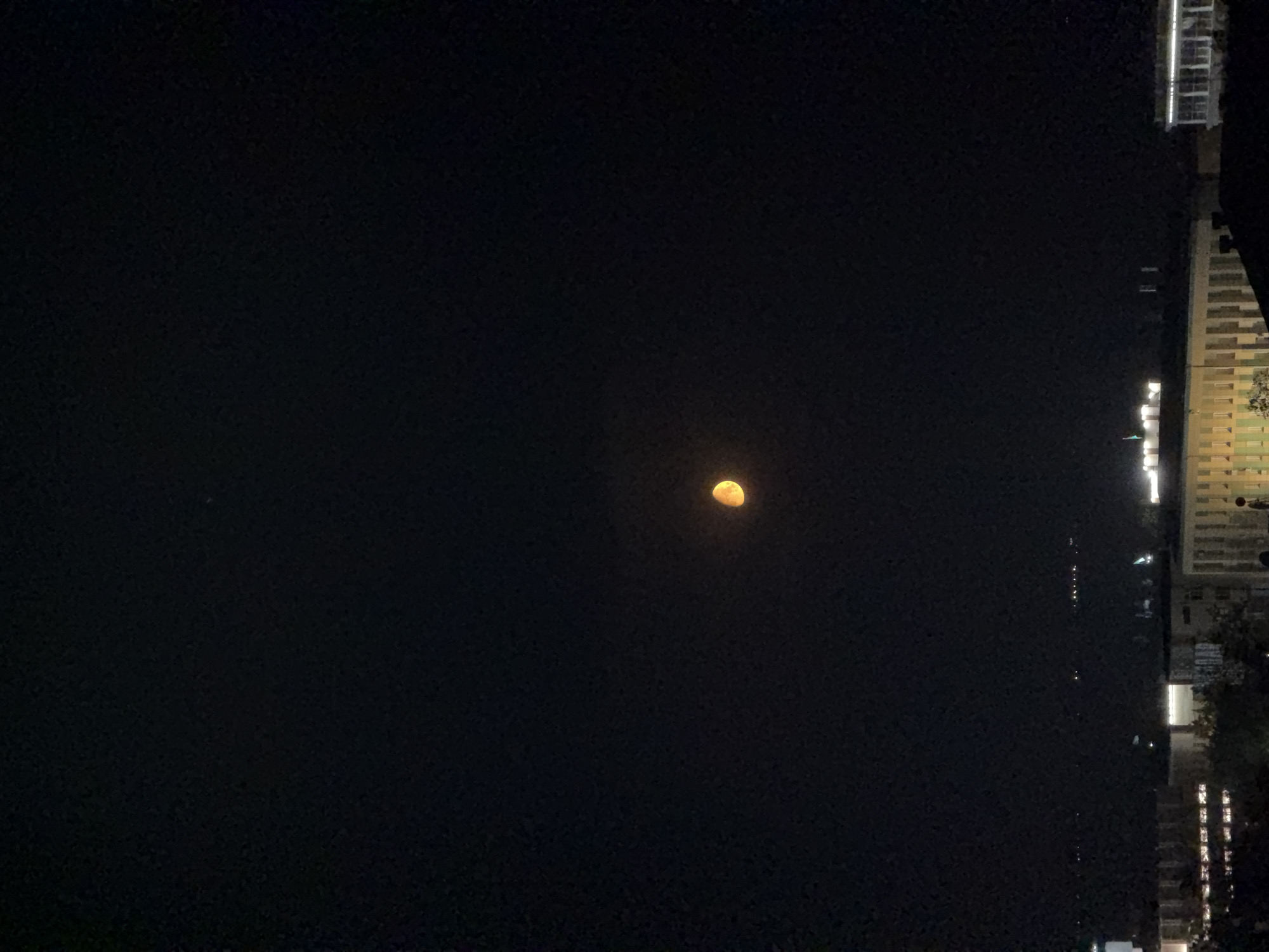A half-illuminated moon hangs in a dark night sky over the city of Pavlodar, Kazakhstan. The moon appears orange-yellow, suggesting it is near the horizon or affected by atmospheric conditions. Below the moon, a cityscape is visible, with illuminated windows and building structures. The lights of distant structures are also present, hinting at the urban environment. The overall impression is of a quiet night in a city.