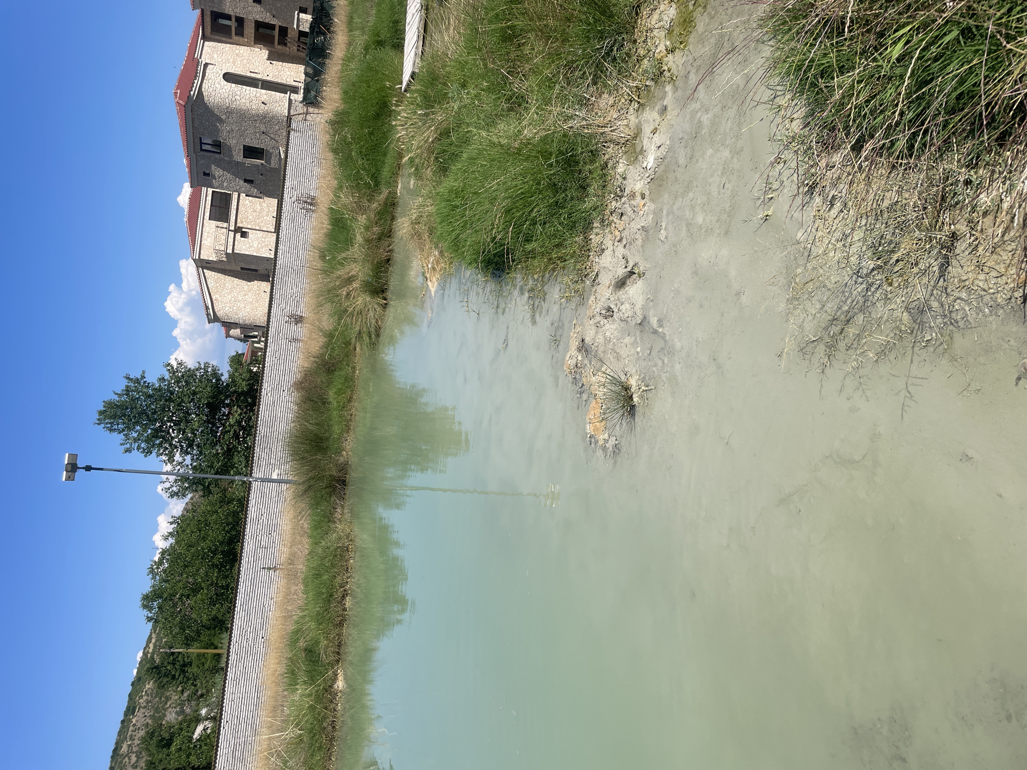 This image depicts a sunlit outdoor scene in General Todorov, Bulgaria, featuring a body of still, milky green-blue water in the foreground, bordered by a muddy bank and various grasses. The water's surface reflects the clear blue sky, distant trees, and a tall metal light pole equipped with two rectangular luminaires, positioned on the left bank.
The immediate shoreline consists of light greyish-tan sediment, transitioning into areas of dense, vibrant green grass and some drier, lighter-colored reeds and scrub. A small, light-colored wooden structure, possibly a platform or part of a boardwalk, is partially visible on the far right bank amidst the vegetation.
In the midground, a low, light-colored horizontal wall, constructed from what appears to be stacked concrete or stone panels, extends across the scene. Behind this wall, to the right, are several multi-story buildings with light beige or pale stone facades and distinct red tile roofs. These structures feature architectural elements such as rounded sections, arched windows, and balconies with dark railings. One building on the far right appears to be under construction or renovation, with exposed structural elements and green netting visible.
The background on the left reveals a vegetated hill or mountain under a clear blue sky with scattered white cumulus clouds. Numerous green trees are visible both behind the wall and along the distant hillside. No people or animals are present in the scene.