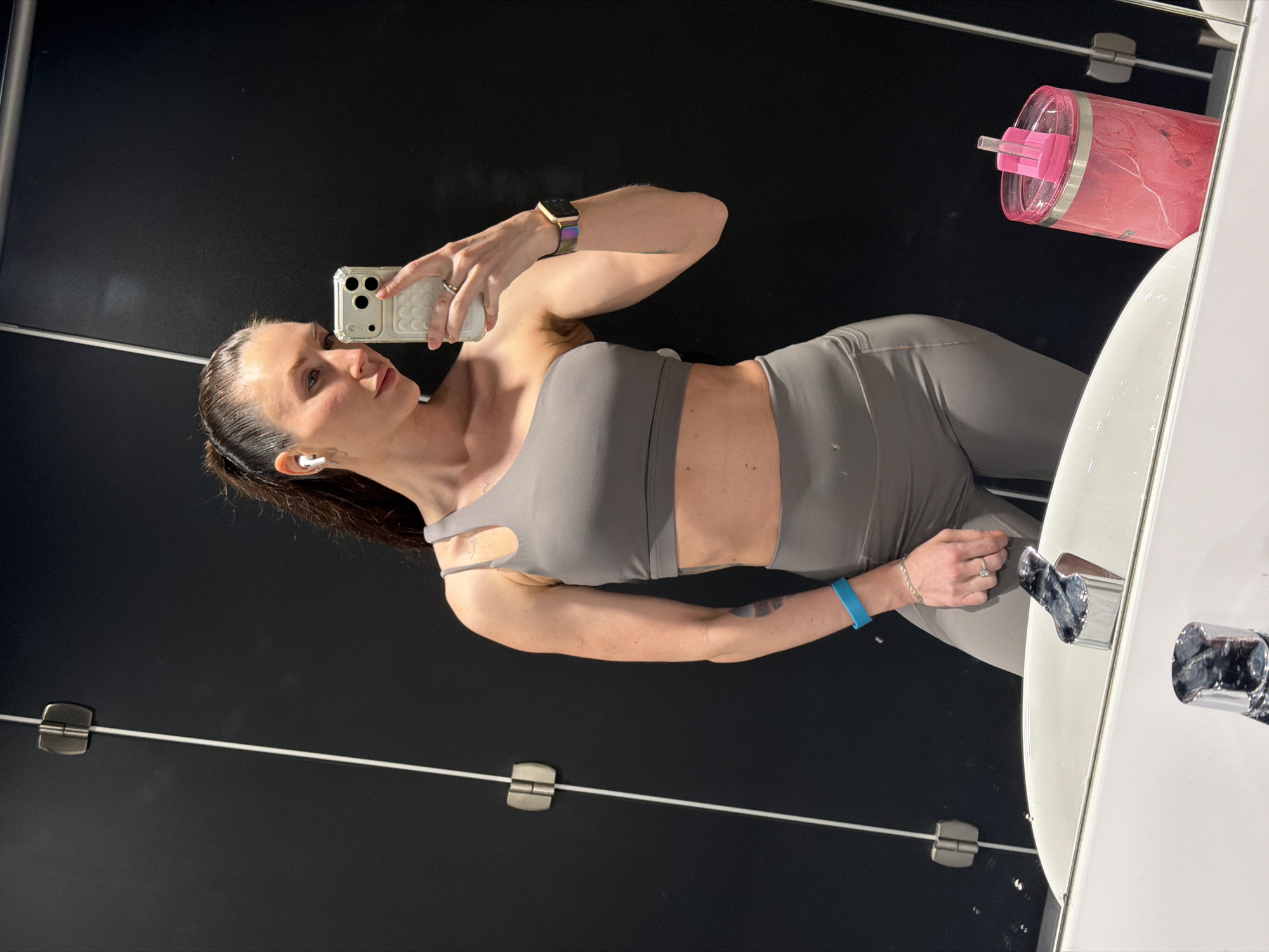 A woman with brown hair pulled back into a ponytail is taking a selfie in what appears to be a public restroom. She is wearing a grey workout set with a sports bra and leggings. She has an Apple watch on her left wrist, a blue bracelet on her right wrist and a phone in her right hand. There is a pink water bottle on a white counter top in front of the sink, and black walls with metal hardware behind her. The location is the city of Perm, Russia.