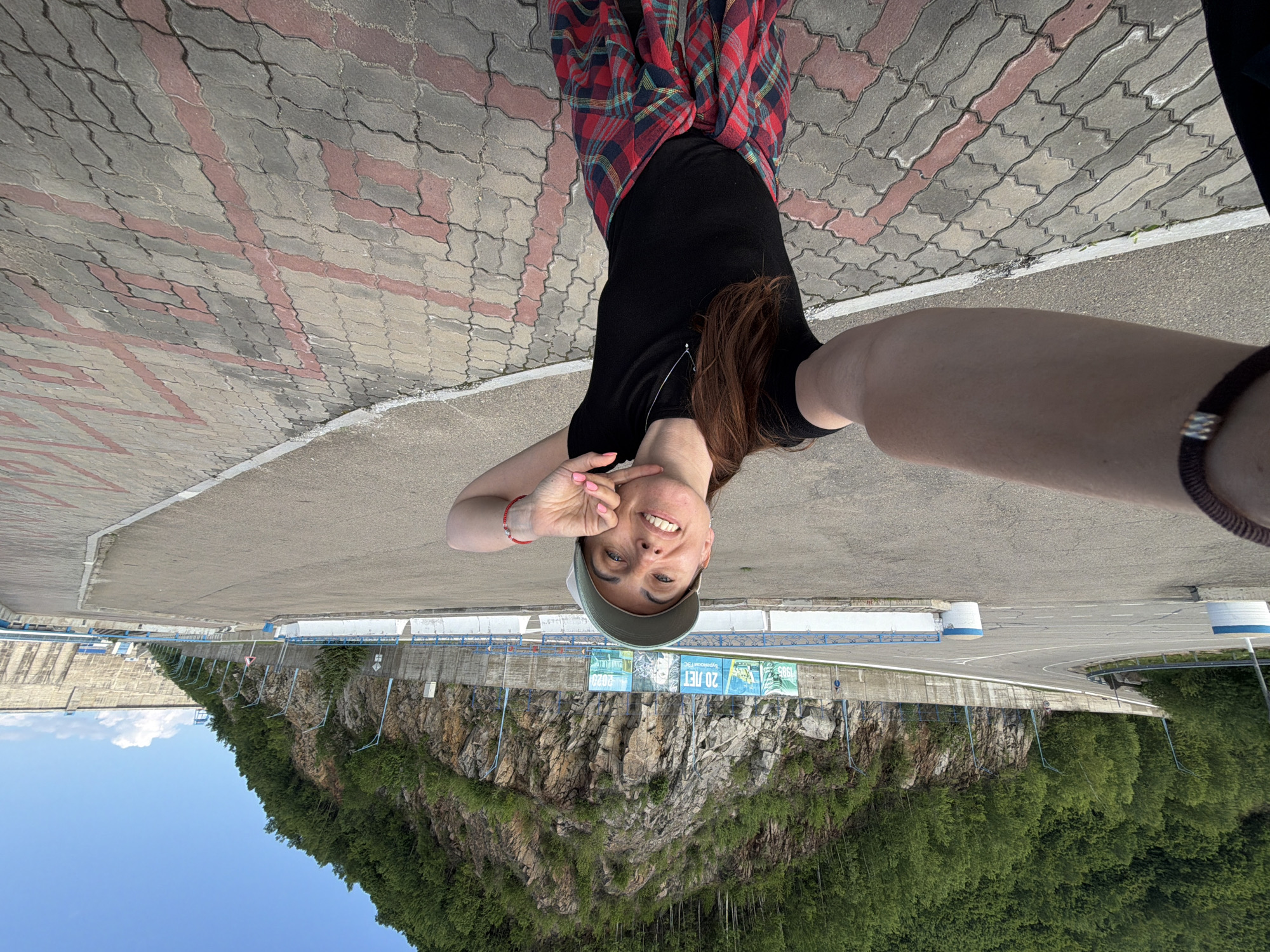 A woman wearing a green baseball cap and a black t-shirt is taking a selfie. She has a red and blue plaid shirt tied around her waist. She is standing on a paved area with red and gray interlocking stones in front of a concrete dam and a rocky hillside covered in trees. In the background, banners are hung on a fence. The sky is clear blue with a few white clouds. The image appears to have been taken in the city of Talakan, Russia.