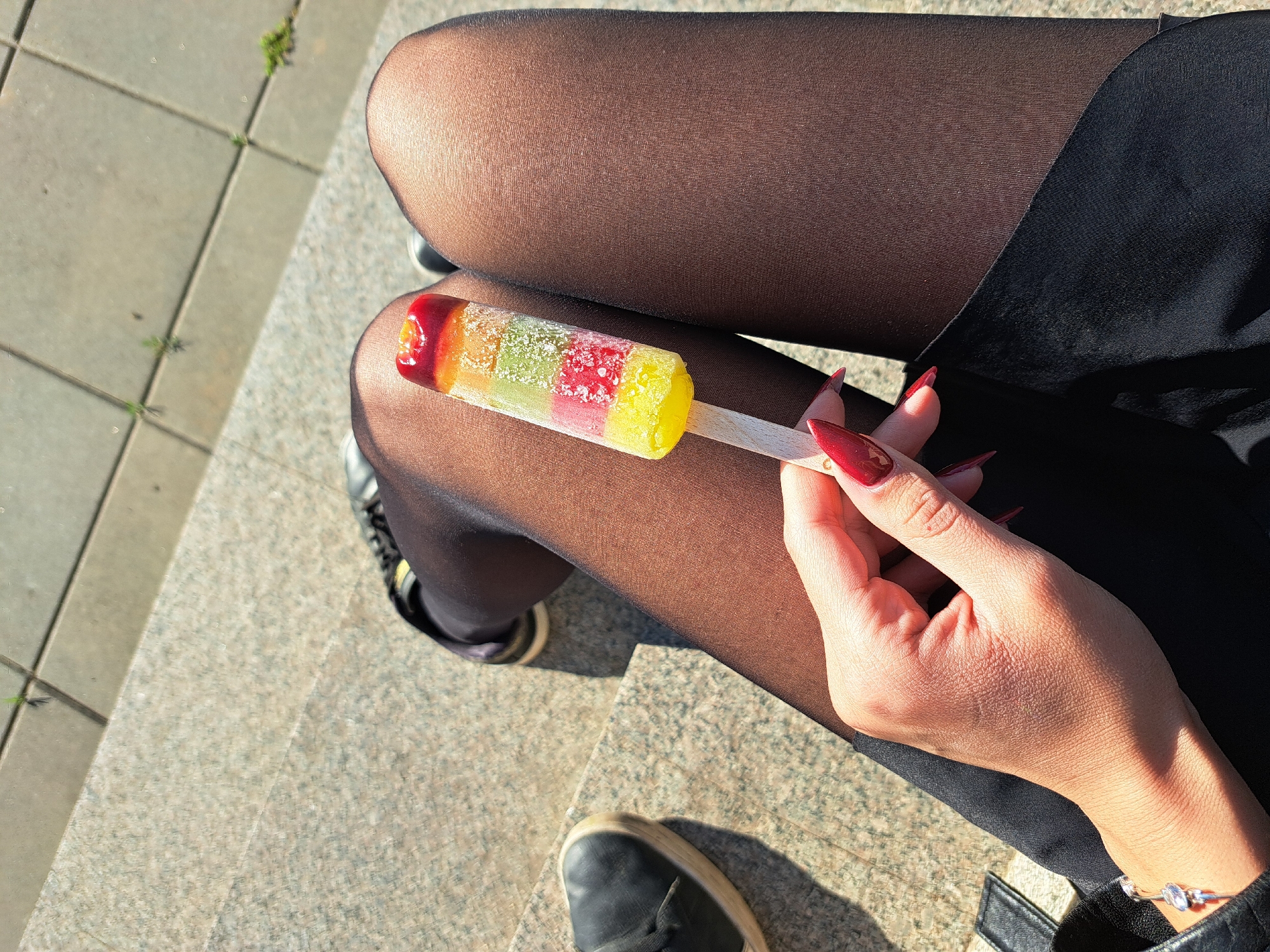 A woman sits on granite steps wearing black tights and a black skirt. She holds a multicolored popsicle with red, green, and yellow sections in her right hand, which is adorned with long, red fingernails and a silver bracelet. Part of her black sneakers are visible. The location may be Negovan, Bulgaria, but there is not enough information to make that deduction.