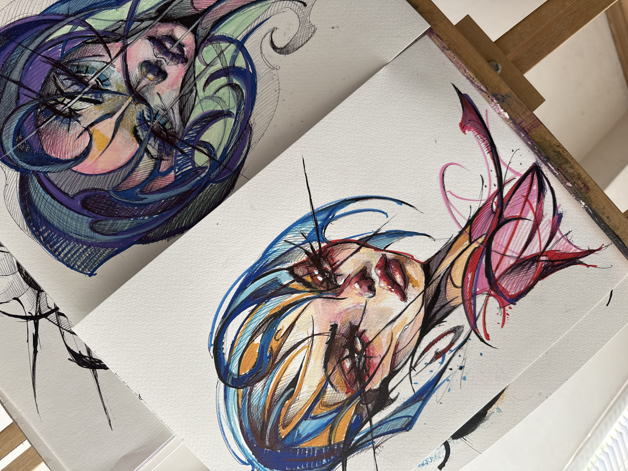 The image shows two watercolor and ink drawings of stylized faces, presumably portraits, arranged on an artist's easel.
The drawing on the left features cool tones of blue, purple, and green. The face is pale pink with black ink details delineating features and creating depth. Swirling lines of color encompass the face.
The drawing on the right features warmer tones of blue, orange, red, and pink. The face is painted in pink and orange with similar black ink details. This face is also surrounded by swirling lines of color.
The drawings are positioned on white paper. The easel is wooden and shows some paint splatters, indicating active use. The background is out of focus and light, suggesting an interior space.
There is no indication that the image was taken in Sofia, Bulgaria.