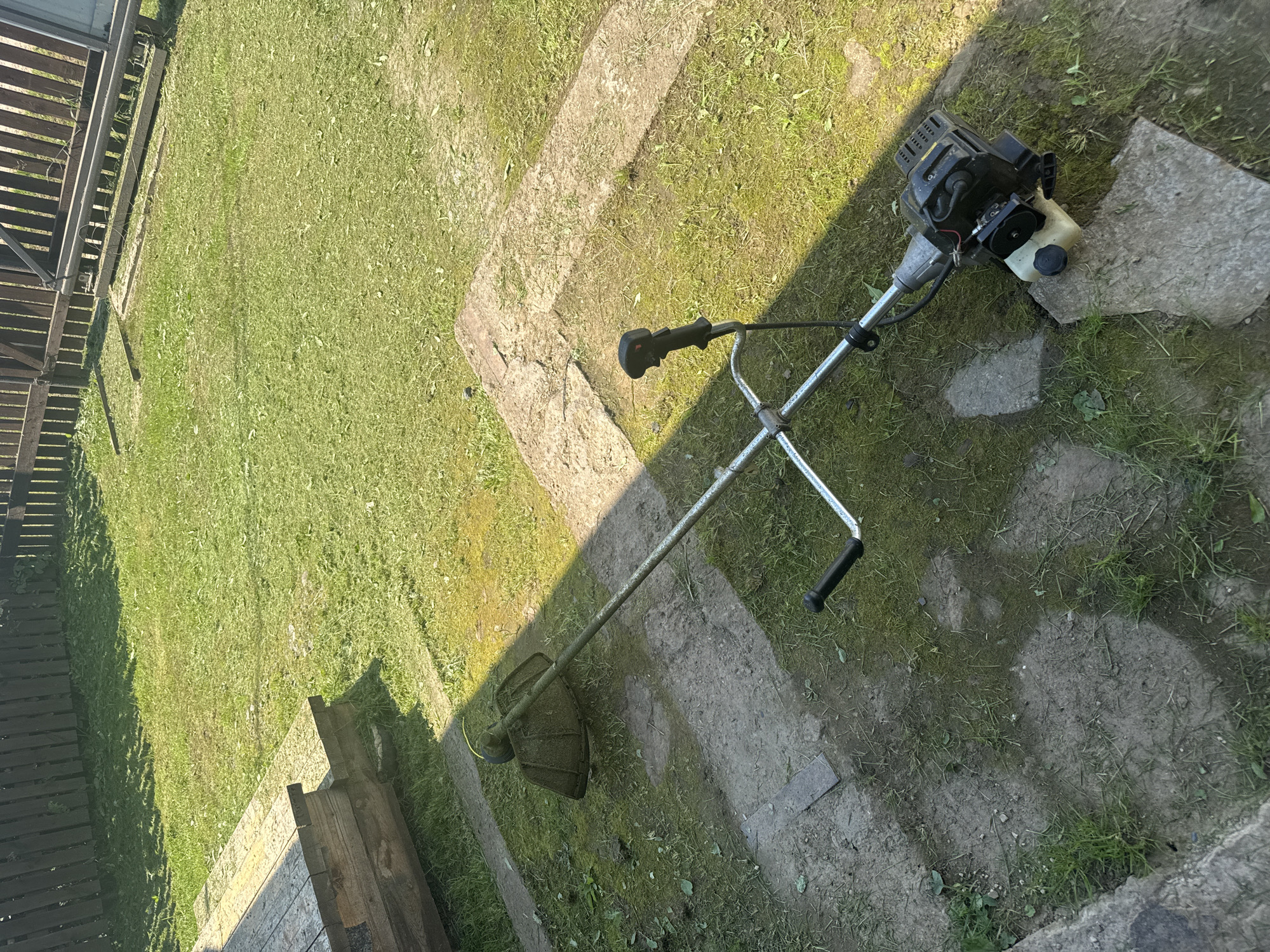A string trimmer rests on a patch of grass and concrete in an outdoor yard. The trimmer is silver and black with a black handle and a yellow string spool. It is positioned diagonally across the frame, with the cutting head on the left and the engine on the right. The yard is partially covered in grass and scattered with some patchy dirt areas and concrete slabs. In the background, a wooden fence runs along the edge of the yard, and beyond that, more grass. The lighting suggests it is daytime, with distinct shadows cast across the scene, indicating sunlight. There are no people visible in the image.