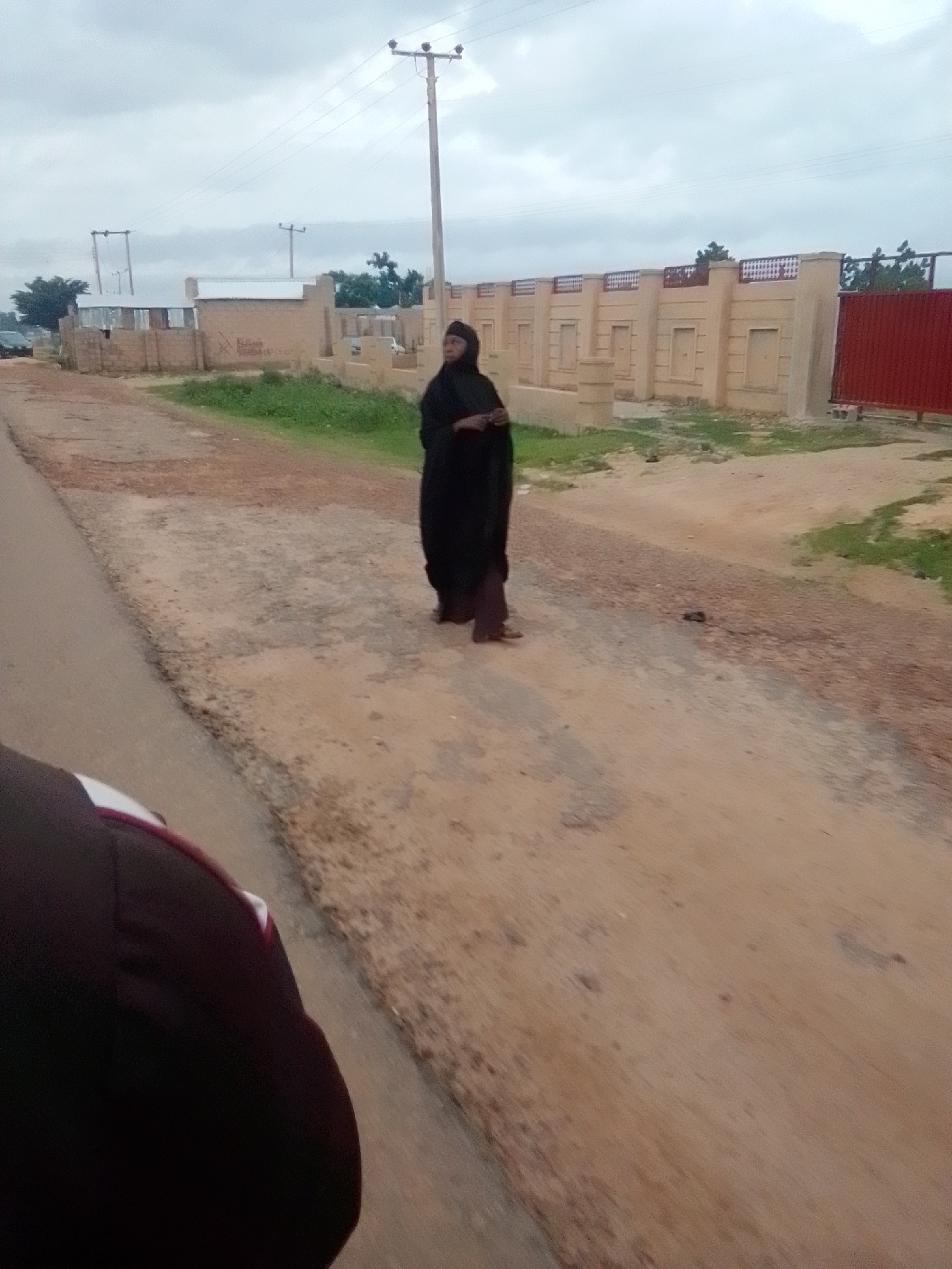 A woman wearing a black hijab and abaya stands on a dirt road in front of a tan-colored building with decorative elements. The building has a metal fence and a red gate. Power lines and a utility pole are visible in the background. The sky is overcast. The scene appears to be in Gombe, Nigeria.