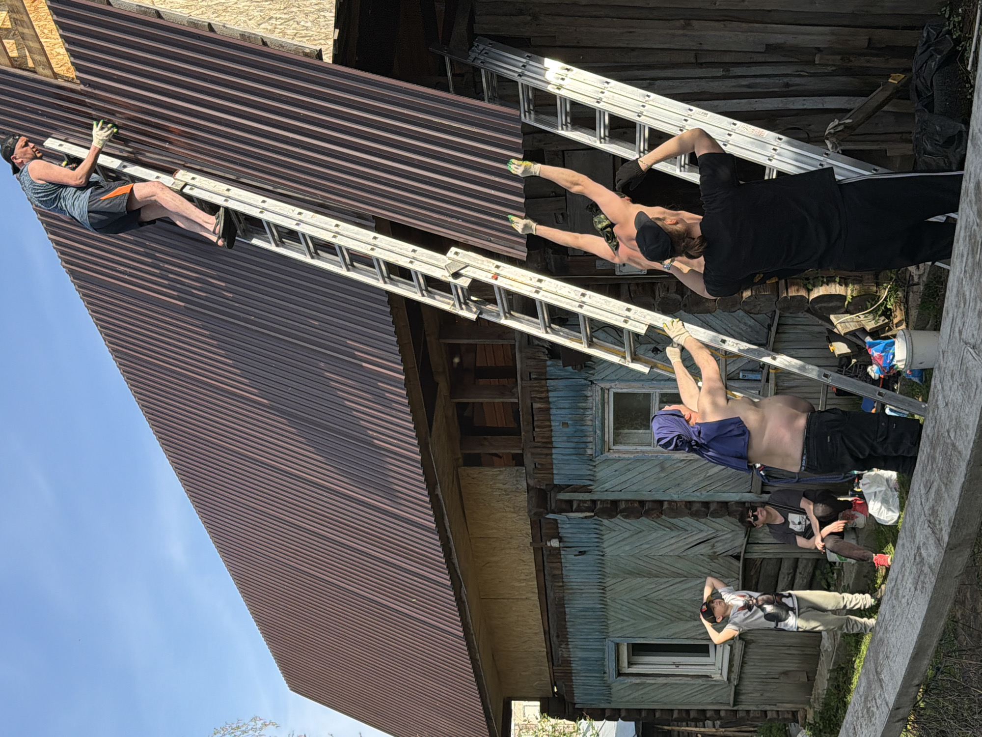 The image depicts a group of people working on a roof in a rural or semi-rural setting, possibly in or near Perm, Russia.
There are six individuals visible in the image. One person is on the roof, presumably working on the corrugated metal roofing material. This person is wearing a tank top, shorts, work gloves and a baseball cap. Another person is standing on a ladder handing something to the person on the roof. The other people are standing on the ground, possibly assisting. One is wearing black pants, a black shirt and a baseball cap. One is shirtless and wearing a blue scarf or piece of cloth. One is sitting and another is standing.
The house appears to be a rustic wooden structure with weathered blue or green siding. The environment includes elements of both construction and nature, with building materials, tools, and greenery visible. Ladders are set up against the building, indicating ongoing construction or repair work.
The sky is visible in the background, suggesting it is daytime.