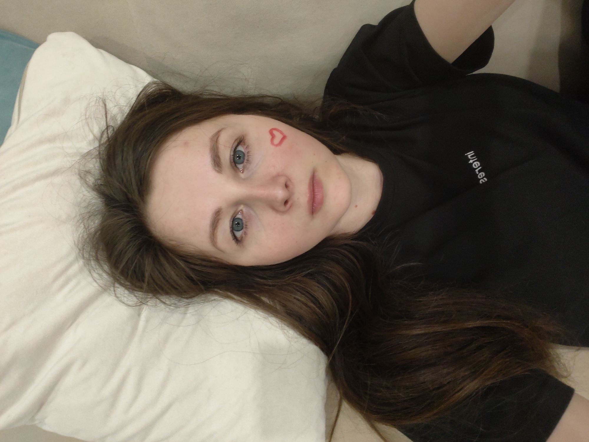 A person lies on a light-colored pillow and sofa. The person has light skin, brown hair, and blue eyes. There is a red heart drawn on the person's cheek. The person is wearing a black shirt with white text. The location is Unknown, Unknown.