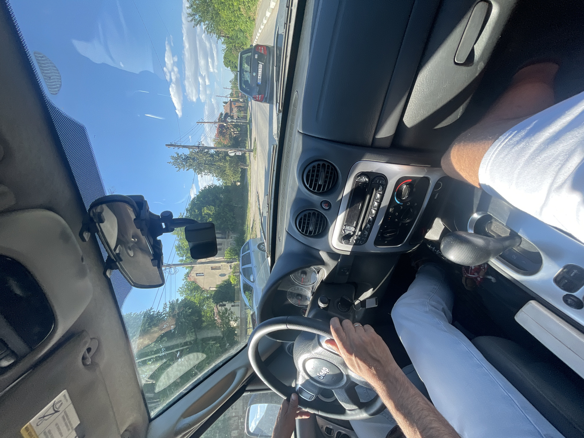The image captures the interior view of a Jeep Liberty, likely in the city of Enitsa, Bulgaria, as indicated by the license plate of the car ahead. The perspective is from the passenger seat, looking forward and slightly to the left. Sunlight streams through the windshield, illuminating the dashboard and the hands of the driver on the steering wheel. The driver, wearing light-colored jeans, is operating the vehicle. In the foreground, a person's legs, clad in white shorts, are visible, suggesting they are either the passenger or the driver in a different seating position than implied by the steering wheel.
The dashboard features the characteristic Jeep design, with prominent air vents and a center console housing the radio and climate controls. A rear-view mirror hangs from the windshield, reflecting a portion of the sky and the car's interior. Outside the car, a street scene unfolds. The road is paved, with a sidewalk and trees lining its sides. Houses with tiled roofs and architectural styles common in Bulgarian villages are visible in the background.
Ahead of the Jeep, a dark-colored Skoda Fabia is seen driving on the same road. Its license plate, "OB 4223 BX," is clearly legible and matches the location specified. The sky is bright blue with scattered white clouds, indicating a clear and sunny day. Power lines and poles are also present, typical of a semi-urban or rural environment. The overall impression is of a casual drive through a residential area on a pleasant day.