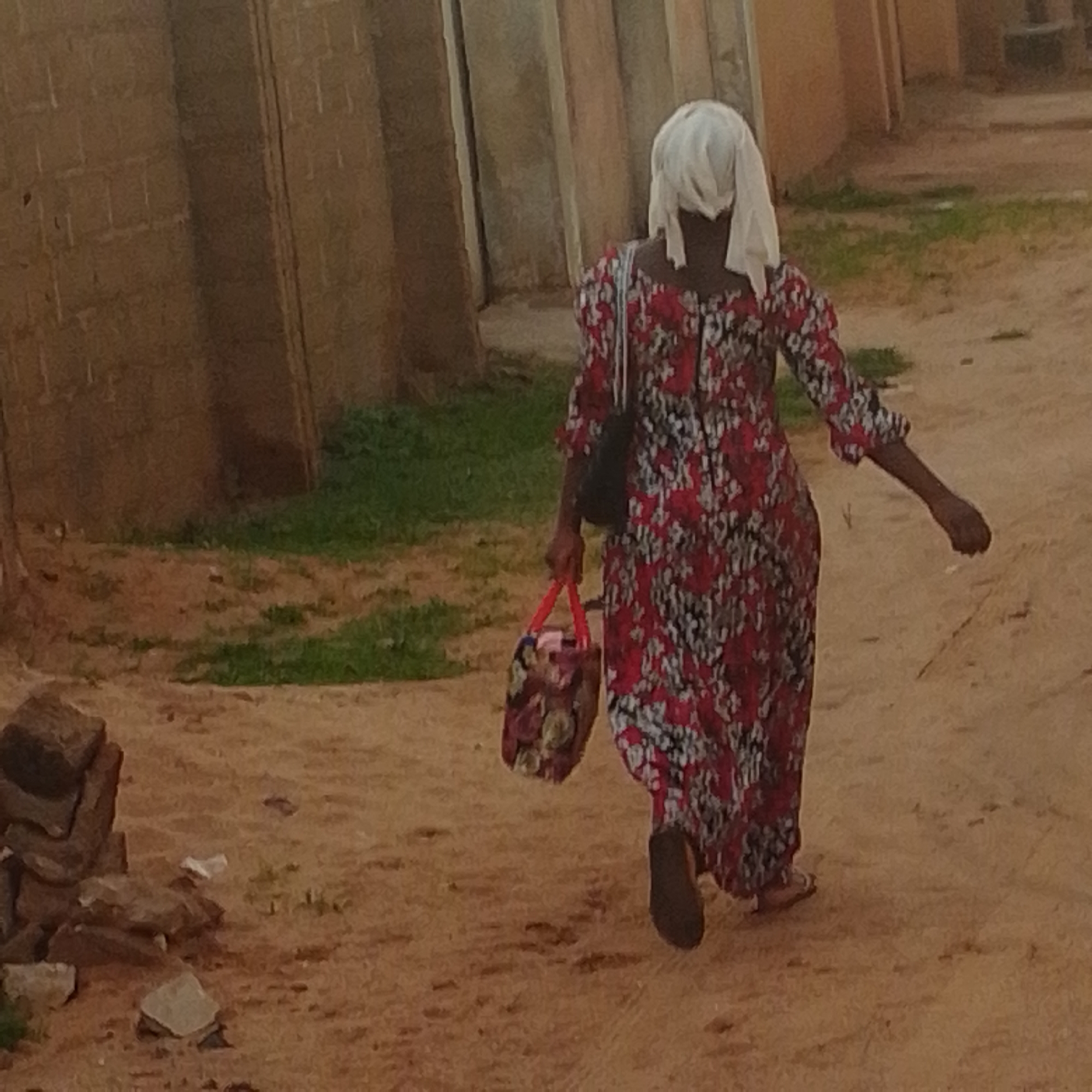 In Tempure, Nigeria, a person wearing a red and white floral patterned dress and a white headscarf walks away from the camera on a dirt path. They are carrying a patterned bag with red handles in their left hand and a dark bag slung over their right shoulder. The background consists of a textured brick wall and a lighter colored wall, with patches of green grass visible. Some rocks are piled on the left side of the foreground. The lighting suggests it is daytime.