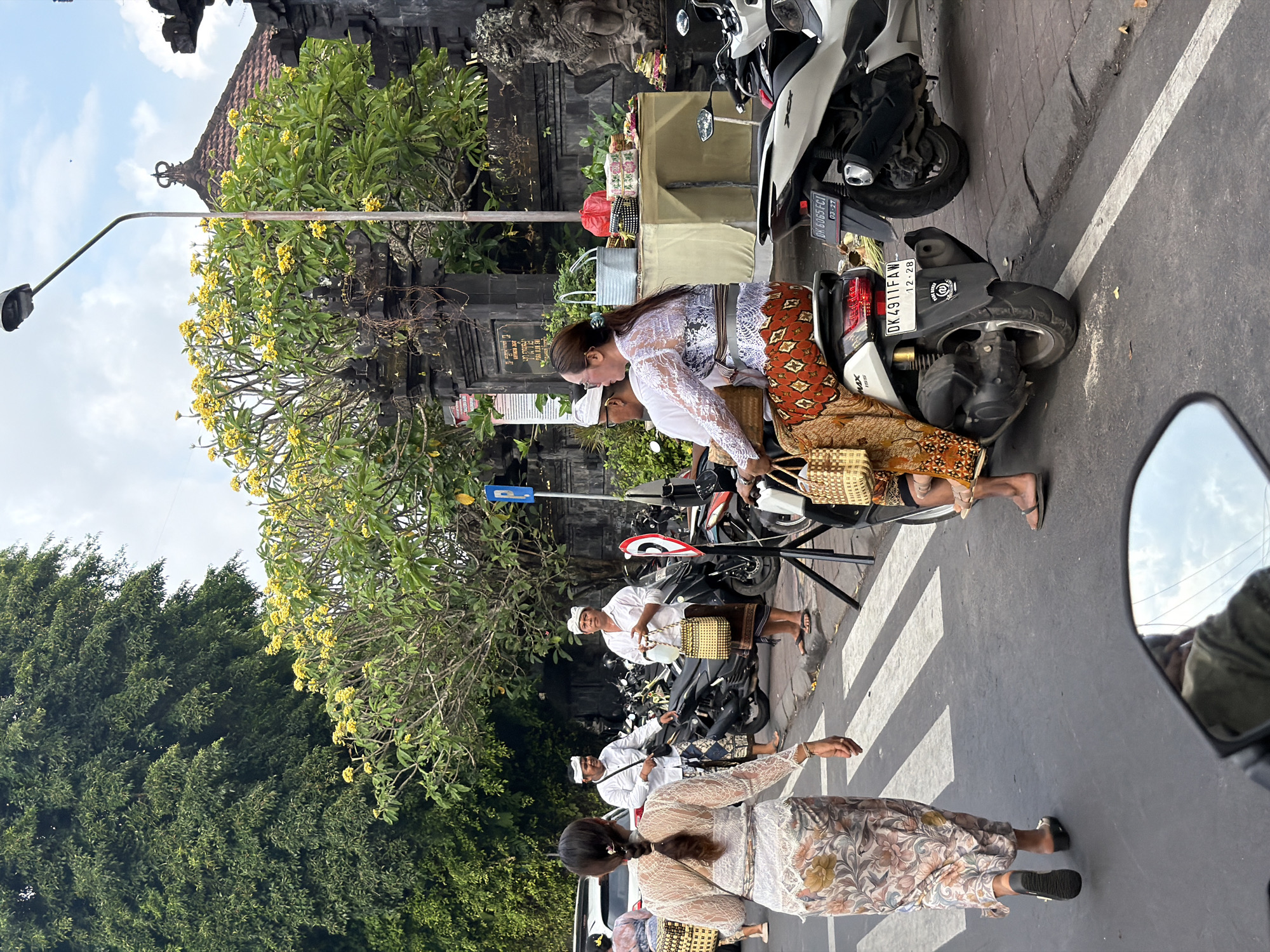 In Kuta Selatan, Indonesia, a street scene unfolds with people in traditional attire on a street. In the foreground, a woman wearing a white lace top and a patterned sarong walks across a pedestrian crossing, carrying a woven bag. Behind her, a couple rides a scooter. The woman is dressed in a similar white lace top and patterned sarong, while the man, wearing a white shirt and sarong, drives. They are accompanied by another woven bag.
To the right, two men in white shirts and sarongs stand on the sidewalk, one holding a woven bag and a clear plastic container, the other looking towards the viewer. Behind them, a variety of motorcycles are parked along the street. A table displays offerings, including woven baskets, red and blue bags, and patterned boxes, suggesting a cultural or religious event. A street light stands tall, and lush green trees with yellow flowers provide shade. The architecture in the background features dark stone structures with ornate details and a tiled roof. The sky is partly cloudy.