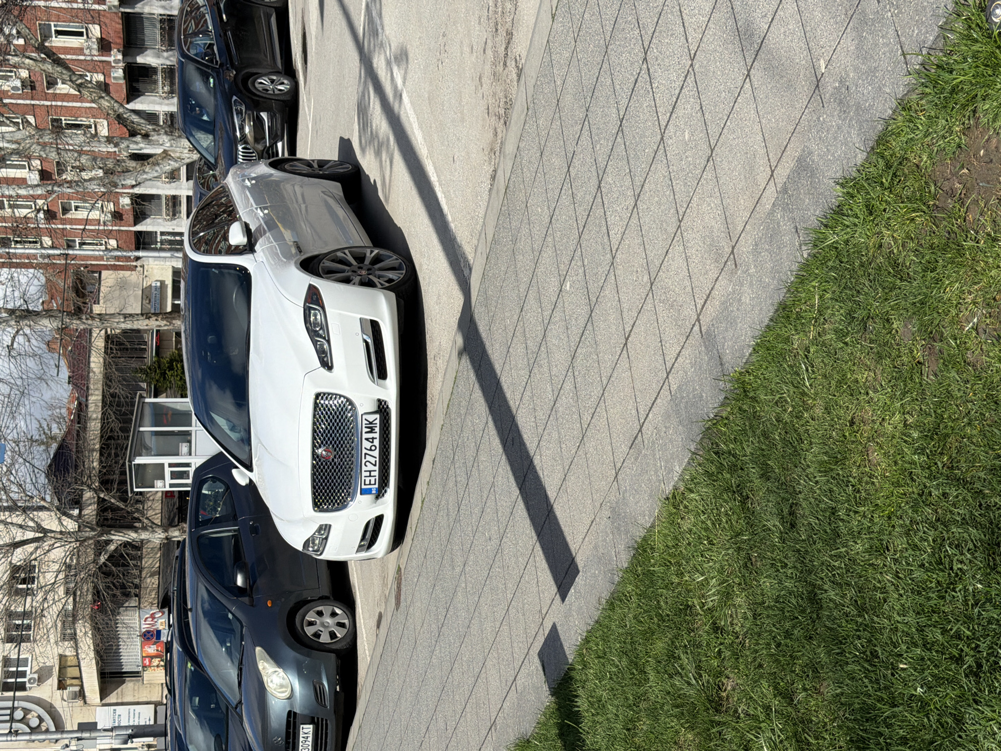 A parked, white Jaguar XJ (license plate EH 2764 MK) sits adjacent to a walkway with light-grey brick paving, next to an area of green grass. Several other cars are parked in the background, behind the Jaguar. In the background, a light-colored building stands next to bare deciduous trees. From the license plate, the car is registered in Bulgaria. From the architectural design of the building, it appears to be in the city of Pleven, Bulgaria.