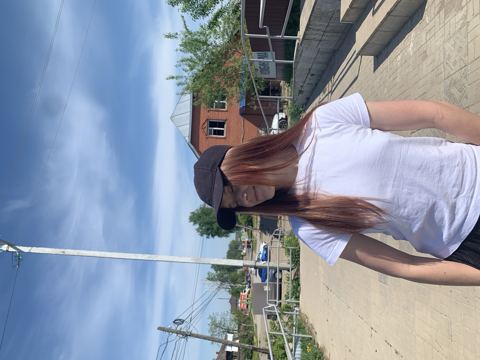 A young woman with long, reddish-brown hair and wearing a black baseball cap and a white T-shirt is standing outside in Фоки, Russia. She is looking down. A paved area is visible beneath her. In the background, there is a building made of red bricks with a gray roof. Street signs, overhead power lines, a blue sky with white clouds, and other buildings can be seen.