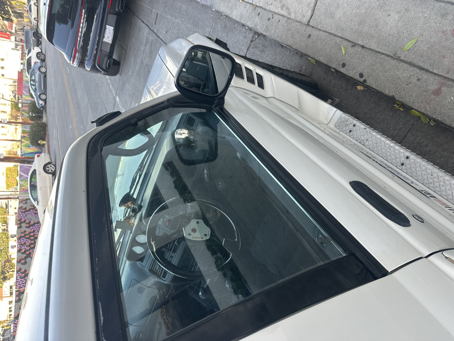 An angled close-up shows the front of a white vehicle, possibly a truck, parked on a city street. The front windshield is visible, with the interior including a steering wheel and dashboard partially seen. The side mirror extends outward. In the background, other parked vehicles, including a black Porsche, line the street alongside buildings and a mural-covered wall. The curb and sidewalk are also partially visible on the right.