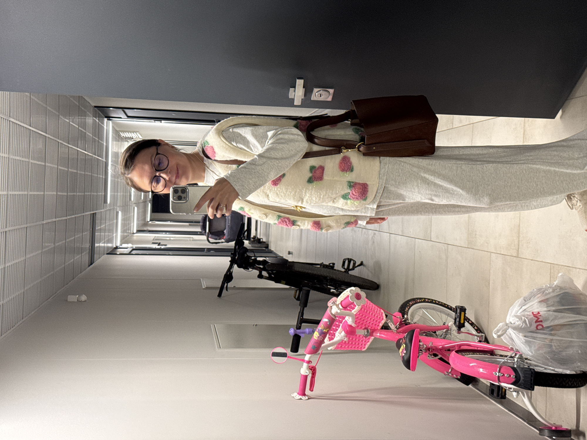 An indoor, vertical-orientation selfie captures a woman standing in a well-lit hallway. She wears glasses, a light-colored long-sleeved top, and a cream-colored vest adorned with pink floral or strawberry-like patterns. A dark brown shoulder bag hangs on her right shoulder. She holds a smartphone, appearing to take the photo, and has a neutral to slight smile. Her lower attire consists of light-colored, possibly grey, relaxed-fit pants.
The setting is a narrow corridor with smooth, light-colored floor and walls, and a dark ceiling. Doors are visible on the right side of the hallway, one of which appears to be black. On the left side of the frame, a textured wall can be seen.
To the woman's right, two bicycles are parked against the wall: a black adult bicycle and a smaller, bright pink child's bicycle equipped with a front basket and a kickstand. A white plastic bag with the text "DMC" is draped over the rear wheel of the pink bicycle. The hallway features artificial overhead lighting. The overall scene suggests a common area or bike storage in a residential building, likely in Izhevsk, Russia, as per the provided context.