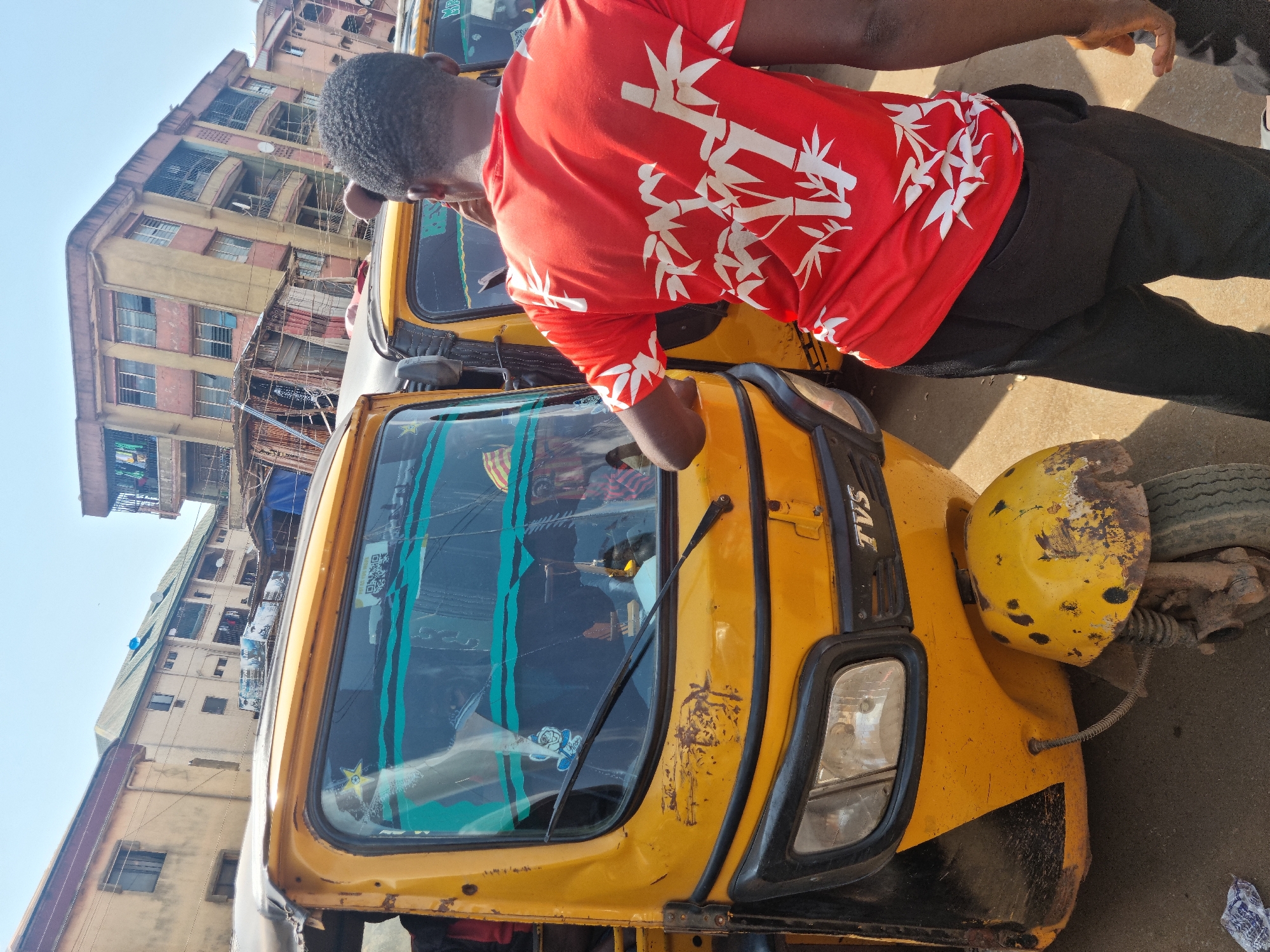 This daytime image, set in Obosi, Nigeria, presents an urban street scene centered around a man and a yellow auto-rickshaw. The perspective is angled, looking down on the subjects.
The main subject is a man with short, dark, curly hair, dressed in a bright red short-sleeved shirt patterned with white bamboo stalks. He also wears plain black pants. He is positioned next to a yellow auto-rickshaw, leaning into its cab with his right arm, suggesting he is either entering the vehicle, preparing to drive, or attending to something inside.
The auto-rickshaw, often referred to as a "keke napep," is predominantly yellow and shows signs of use and age, including some dirt and rust spots, especially on the front fender and around the headlight. The brand name "TVS" is visibly printed in black text on the side near the windshield. The windshield features green decorative stripes on the inside. A single headlight is visible on the right front side of the vehicle.
In the background, several multi-story buildings rise towards a clear, bright sky. These structures appear to be residential or mixed-use, characterized by multiple windows and balconies, and they exhibit an aged aesthetic typical of urban environments. The bright sunlight casts distinct shadows on the ground, indicating clear weather conditions. The overall setting depicts a busy, practical moment in a Nigerian town.