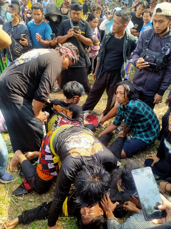 An outdoor scene in Tawangsari, Indonesia, depicts a crowded public gathering where several individuals are on the ground, seemingly in a state of trance or performance, surrounded by a large group of onlookers. In the foreground, two men in black long-sleeved shirts with yellow graphic designs on their backs are on the dirt and sparse grass. One man lies prone on his stomach, head turned left, eyes closed. Another man, also in a similar black shirt, kneels over him, placing his left hand near the first man's head. A third, smaller individual, with blue-dyed hair and red facial marks, also in a black graphic shirt, kneels nearby, possibly holding a decorated, horse-like prop, suggestive of a `Kuda Lumping` performance.
Attending to these individuals are several people: A man in a red and white headband and black shirt bends over the smaller figure. Another man in a blue and black plaid shirt sits on the ground, holding and manipulating the leg of the prone man. Surrounding this central activity is a dense crowd of men, women, and children observing the scene. Many onlookers are holding smartphones, with some actively recording or photographing the event. Notable onlookers include a man in a black shirt and traditional black headwear (udeng) looking at his phone, another man in a dark blue hooded jacket with a chest rig also holding a phone, and a woman in a black hoodie wearing a white face mask. The environment is sunlit, with uneven ground and scattered natural vegetation.