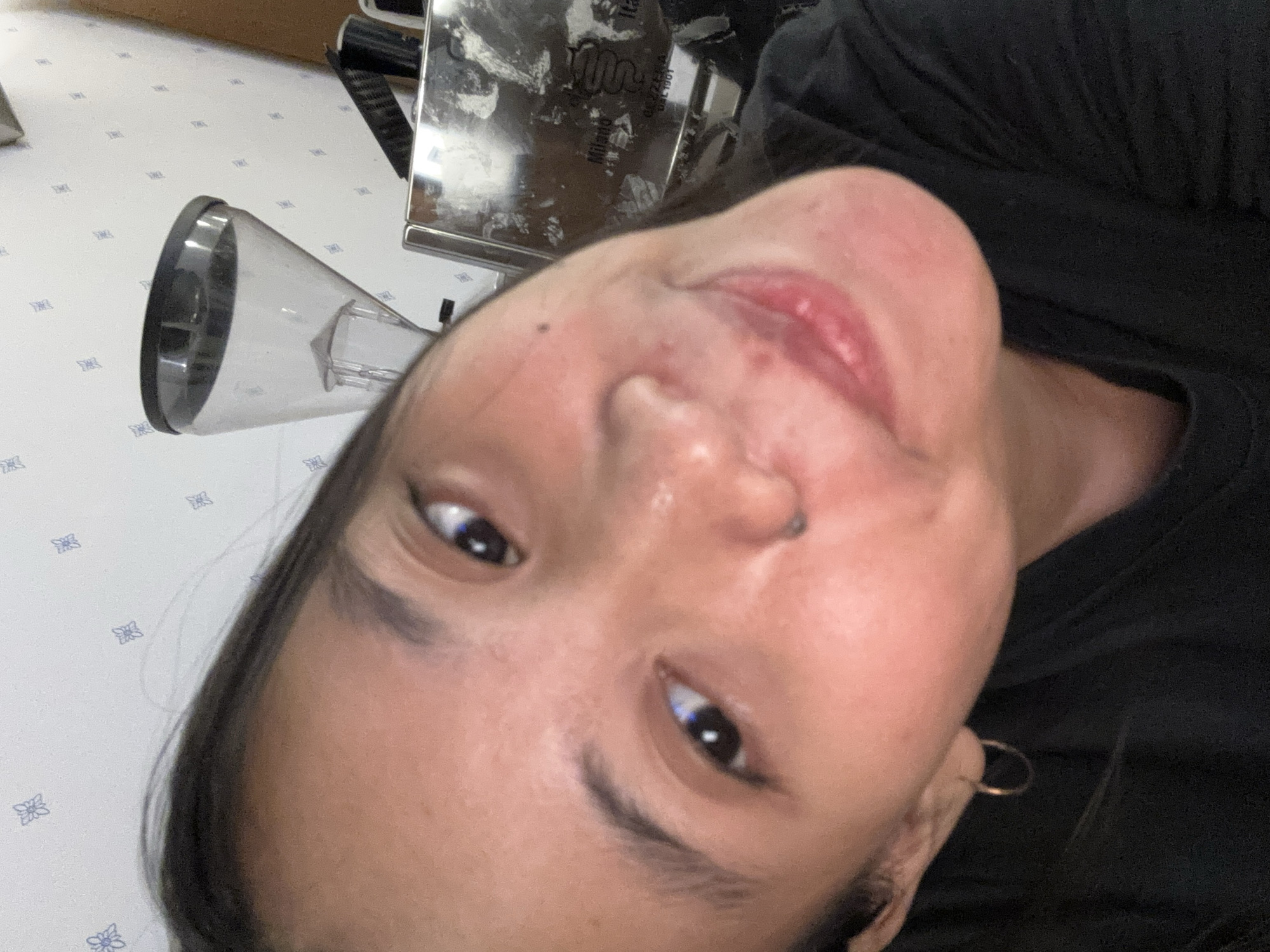 In this close-up selfie, a young woman is positioned slightly off-center, her face angled towards the camera. Her dark hair frames her face, and she is wearing a simple black t-shirt. A small hoop earring is visible on her left ear. In the background, a coffee grinder with a transparent hopper sits on a metallic surface, possibly a coffee machine. The wall behind the subjects is adorned with a subtle, repeating pattern of light blue floral motifs. The lighting suggests an indoor setting, likely a domestic environment. The overall impression is that of a casual, personal moment captured for a photograph. The scene is not specifically identifiable to a particular location within South Jakarta, Indonesia, but the presence of a coffee grinder suggests a home or a cafe setting.