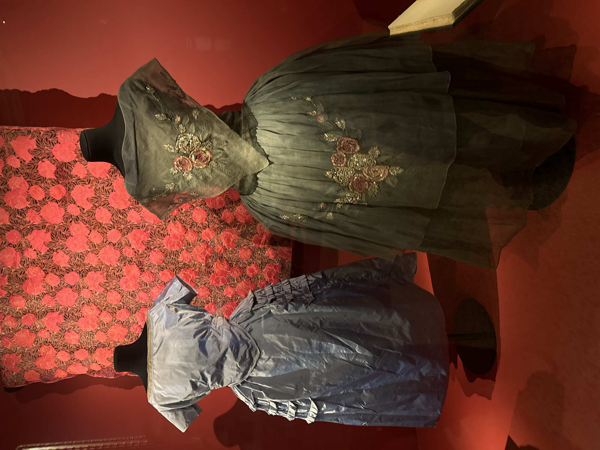The image shows two dresses on display in a museum. The dresses are presented on black mannequins, with one dress appearing blue and the other grey with floral embroidery. The backdrop consists of a red patterned wall. The location is likely in Paris, France, though the specific museum is not identifiable from the image alone.
