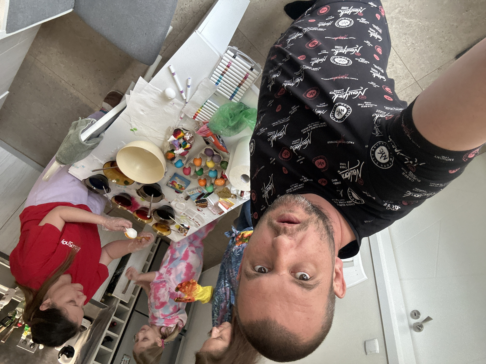 A selfie is taken by a man wearing a black t-shirt with "New York" written all over it. He is standing above a table covered with items related to Easter egg dyeing. A woman in a red shirt and a young girl in a pink tie-dye shirt are dyeing Easter eggs. Bowls of dye, eggs in containers, markers, and paper towels are on the table. The location cannot be determined to be in Sofia, Bulgaria from the provided image.