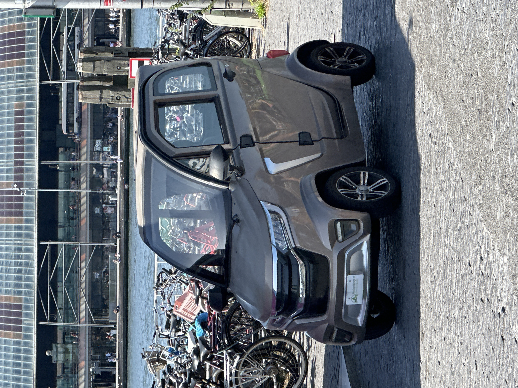 The image shows a small, two-seater car, likely electric, parked on a cobblestone surface. The car is a taupe color with silver accents and black wheels. It has a glass roof and doors. In the background, a body of water is visible, with numerous bicycles parked along the embankment. Behind the water and bicycles is a large, modern building with a glass facade. People are visible on a walkway in front of the building, and a bus is seen on an elevated roadway. The scene appears to be a public area in Amsterdam, Netherlands, given the prevalence of bicycles and the architectural style of the building. The car is positioned in the foreground, taking up a significant portion of the frame.