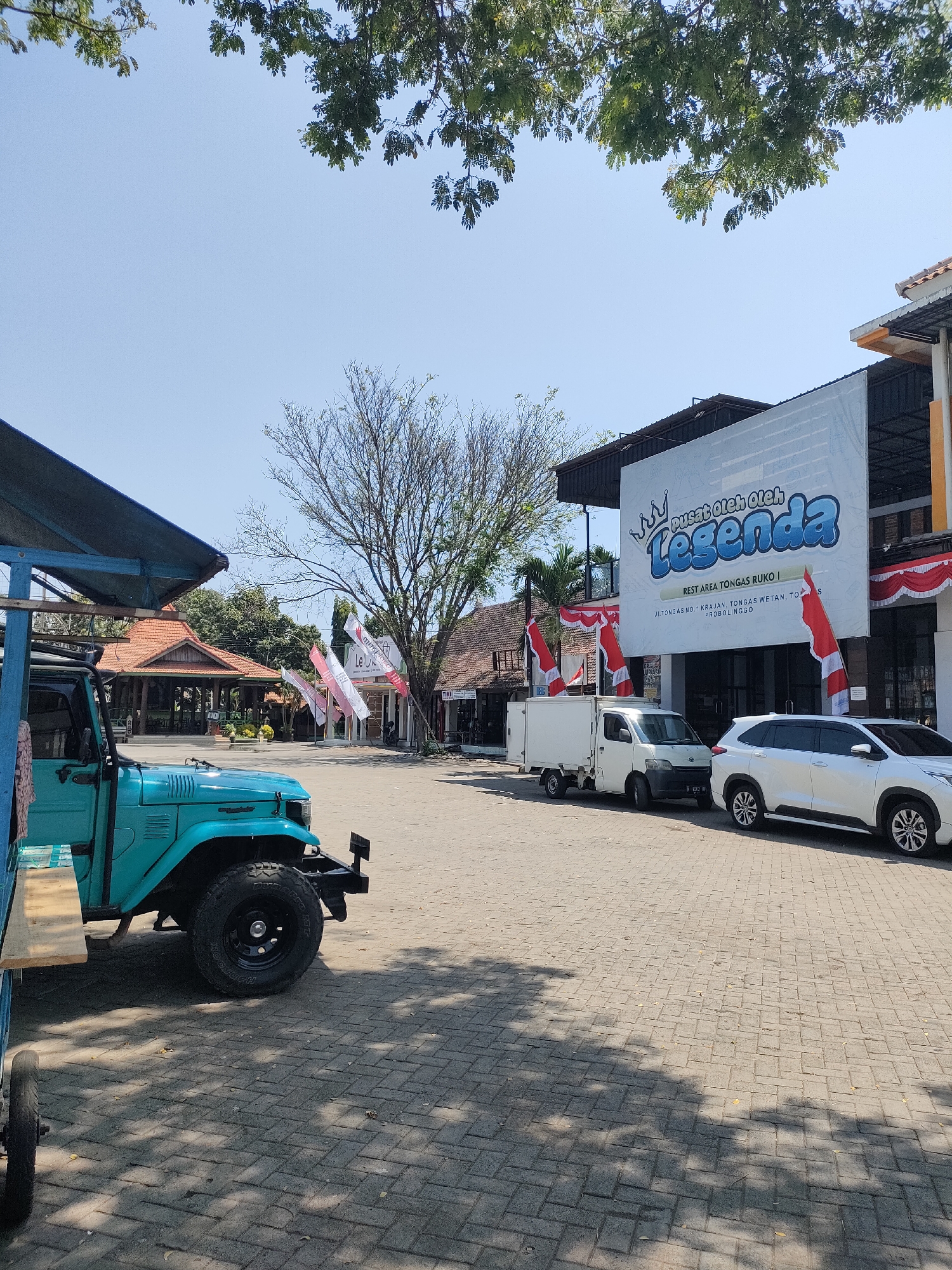 The image shows a street scene in Curahdringu, Indonesia. In the foreground, a turquoise classic Toyota Land Cruiser is parked on a paved area. The vehicle has a spare tire mounted on the rear. To the right of the Land Cruiser, a white delivery van and a white SUV are parked. Behind the vehicles, a large building with a prominent banner that reads "Pusat Oleh Oleh Legenda" (Legend Souvenir Center) is visible. The building has a modern facade with large windows. Several Indonesian flags are displayed in front of the building. In the background, traditional Indonesian-style buildings with terracotta roofs can be seen. Trees with dense foliage are scattered throughout the scene, and the sky is clear and blue. The overall impression is of a busy roadside rest area or shopping complex.