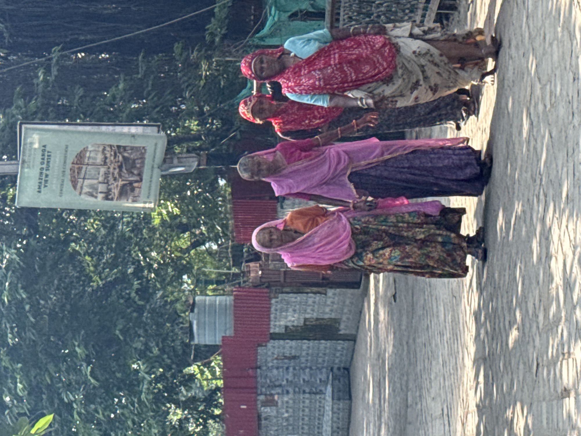 Four women are walking in a line on a paved surface. The woman in the foreground on the left is wearing an orange top and a pink headscarf, with a colorful patterned skirt. The next woman is wearing a purple outfit with a pink headscarf. The third woman, partially obscured, wears a red and black garment with a red headscarf. The fourth woman on the right has on a light blue top and a pink and white patterned headscarf and skirt. In the background, there is a sign attached to a pole that reads "AMAZING GANGA VIEW SUNSET". Behind that, there are trees, a water tank, and buildings with corrugated red roofs. The scene appears to be outdoors in a sunny environment, with shadows cast on the ground. This location is likely near the Ganges River in Rishikesh, India, given the signage and the traditional attire of the women.