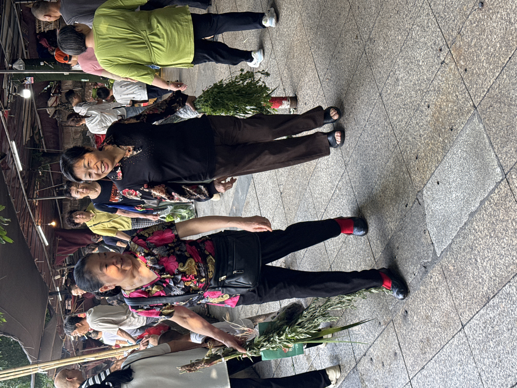 In Jiangjin, China, two women walk side-by-side on a paved outdoor market. The woman on the left, wearing a floral-patterned blouse and black pants with red socks, carries a bundle of greenery and a green bag. The woman on the right, in a black patterned top and brown pants, also carries a bundle of greenery. Both women are smiling and appear to be engaged in conversation. In the background, other people are visible browsing market stalls under awnings. The ground is a mosaic of light gray square tiles.